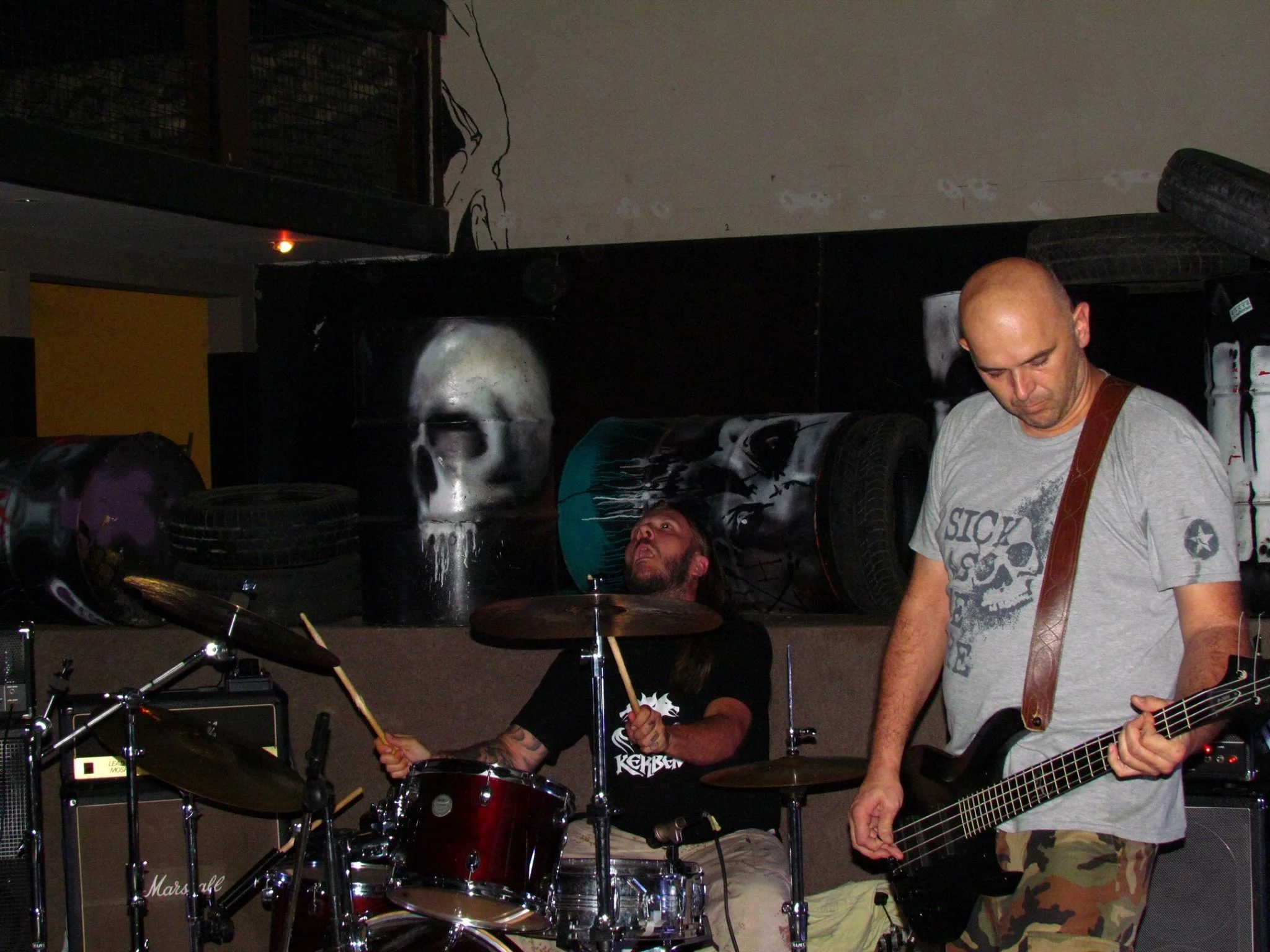 Two musicians perform in a music venue. One is playing drums, and the other is playing an electric guitar. The background features a skull artwork on the wall and several tires stacked on the right side.