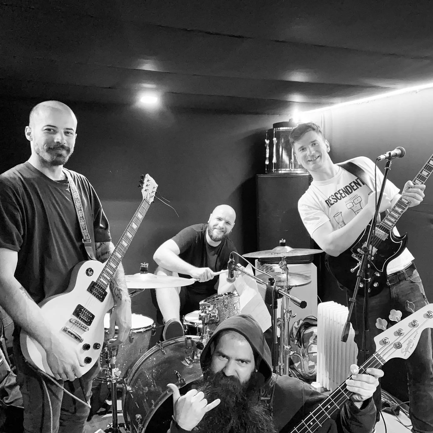 Black and white photo of four musicians in a band playing instruments in a small room. Two guitarists are standing, one drummer is sitting behind a drum set, and a fifth person with long hair and a beard is in the foreground making a hand gesture.