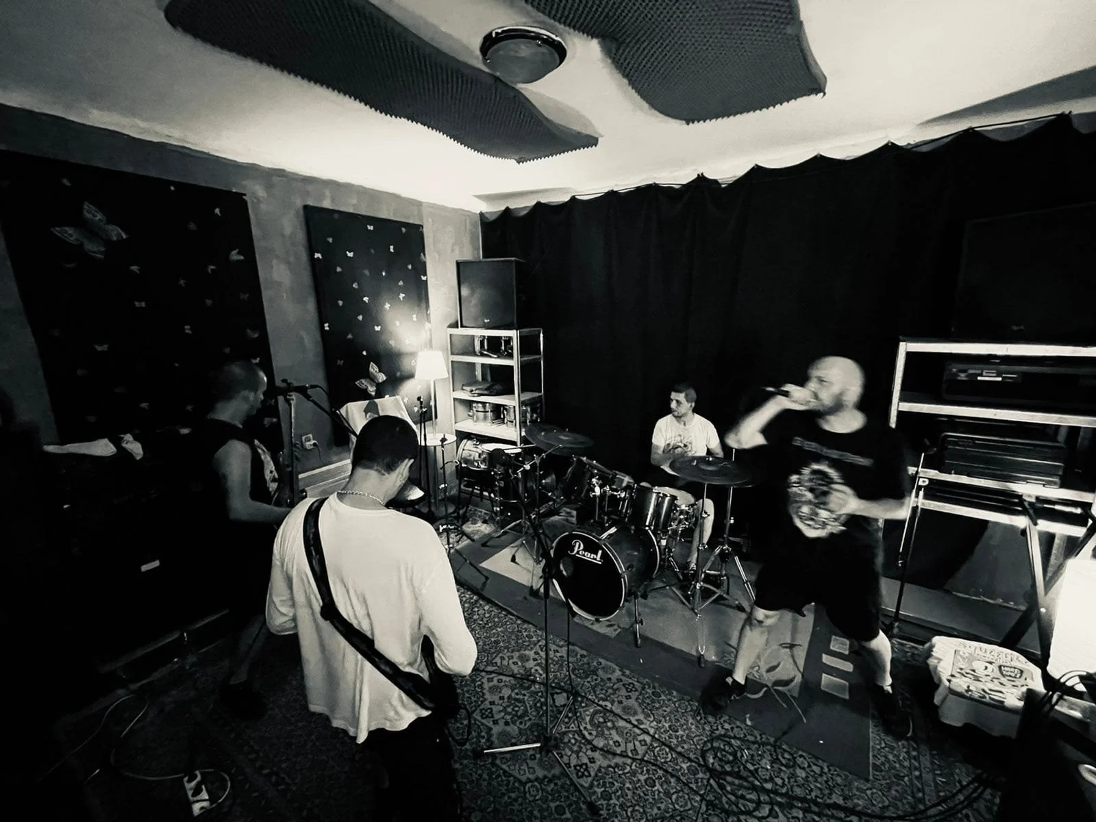 A band of four musicians performing in a small, dimly lit music studio or practice room. One person is playing piano, another is on drums, the third person is standing with a microphone, and the fourth is sitting with their back to the camera.