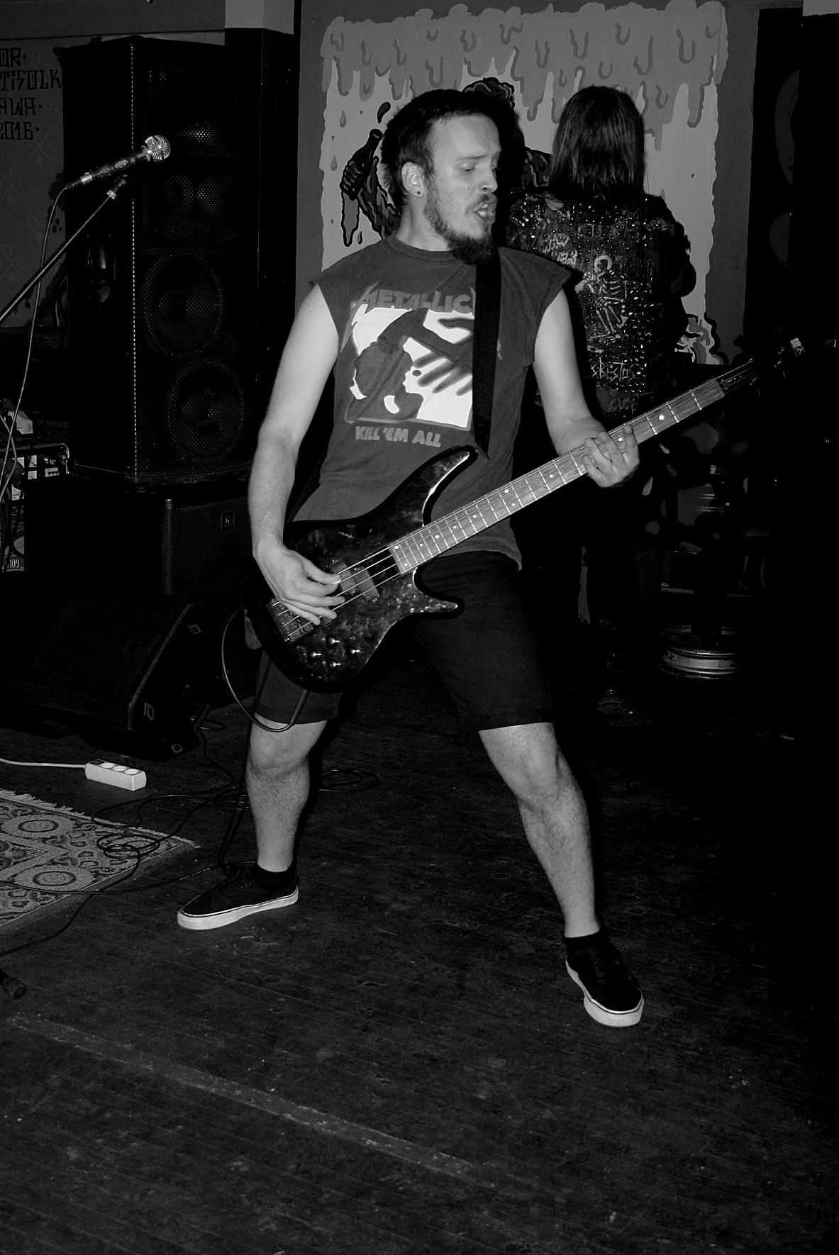 A man playing an electric guitar on stage at a live music venue. He is wearing a sleeveless Metallica shirt, shorts, and sneakers, and appears to be deeply engaged in the performance. There is stage equipment and speakers visible around him, and a wo