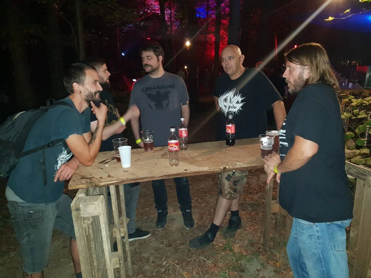 Five men standing and leaning on a wooden table, holding bottles of Coke, socializing outdoors at night in a wooded area with colored lights.