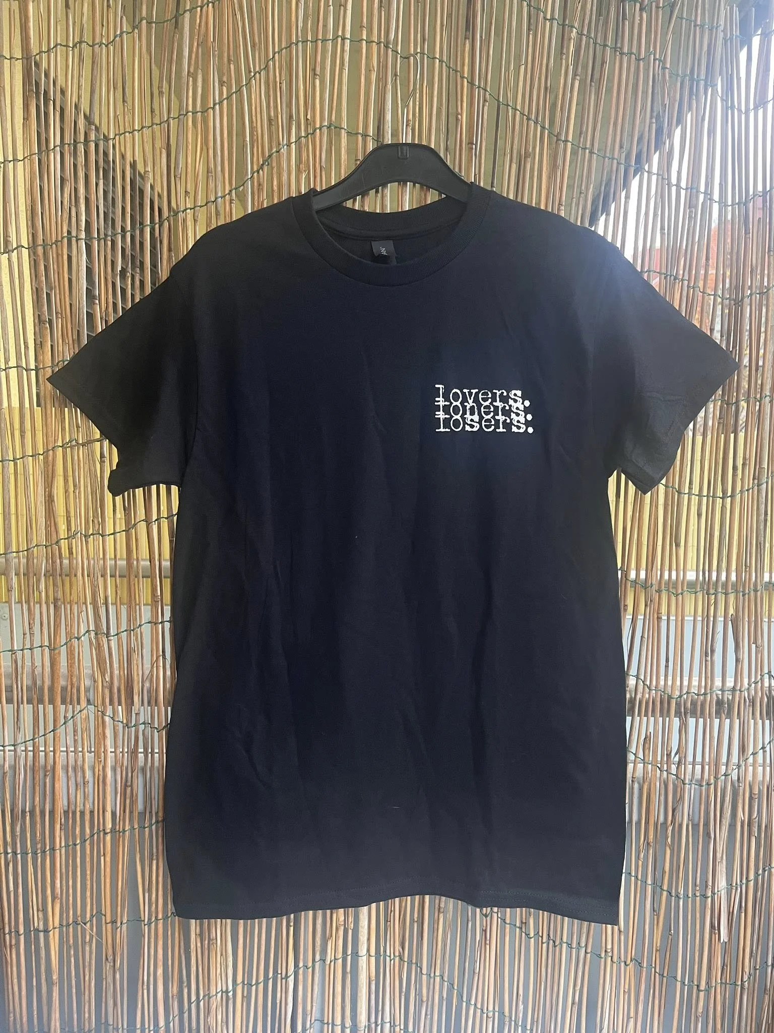 Black T-shirt hanging on a hanger in front of a bamboo backdrop. The shirt has white text on the front that reads 'Lovers. Losers.'