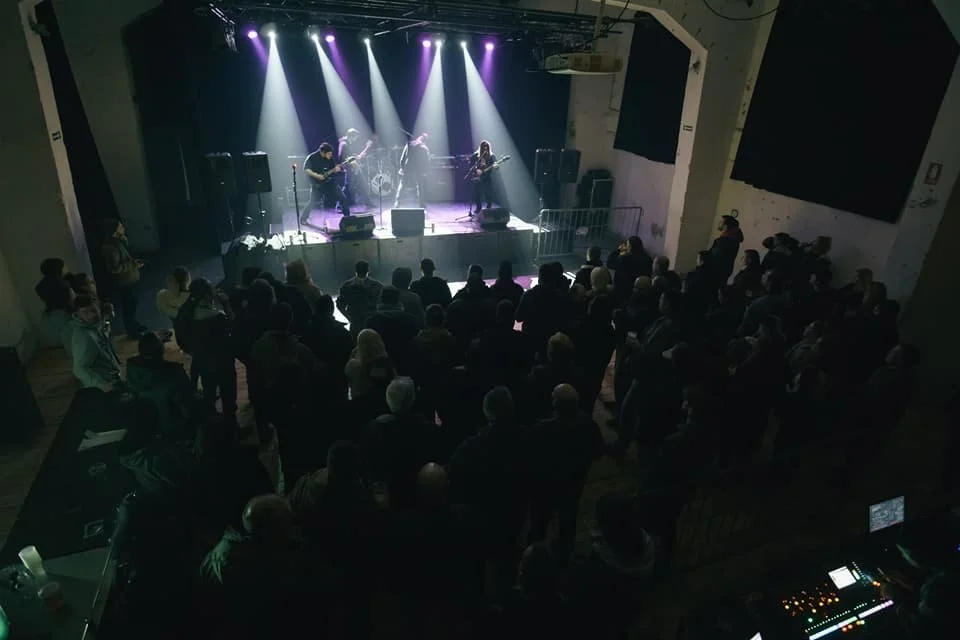 Live performance of a band with three musicians on stage, singing and playing guitar, in front of an audience in a dimly lit concert venue with purple stage lighting.