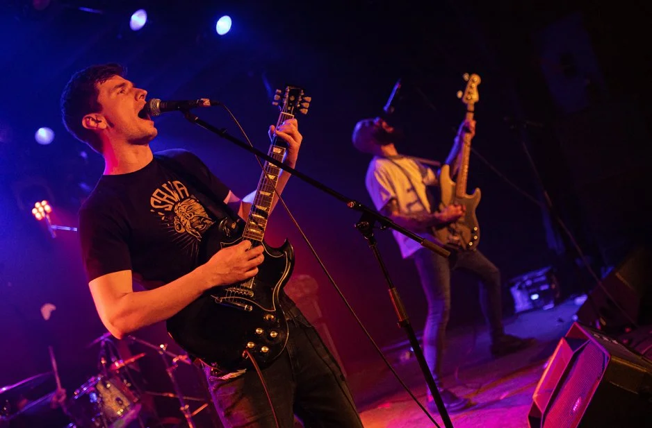 Two musicians performing on stage with colorful lighting; one playing guitar and singing into a microphone, the other playing bass guitar.