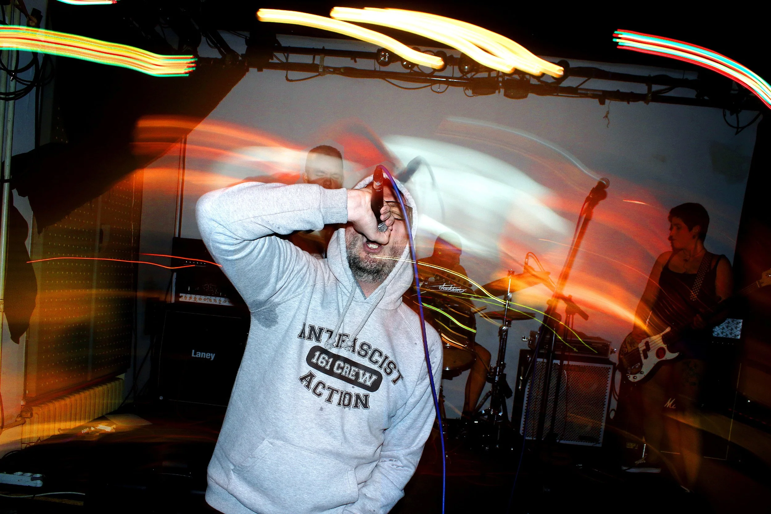 A man singing into a microphone during a live music performance with a band playing instruments in the background, and colorful light streaks across the scene.
