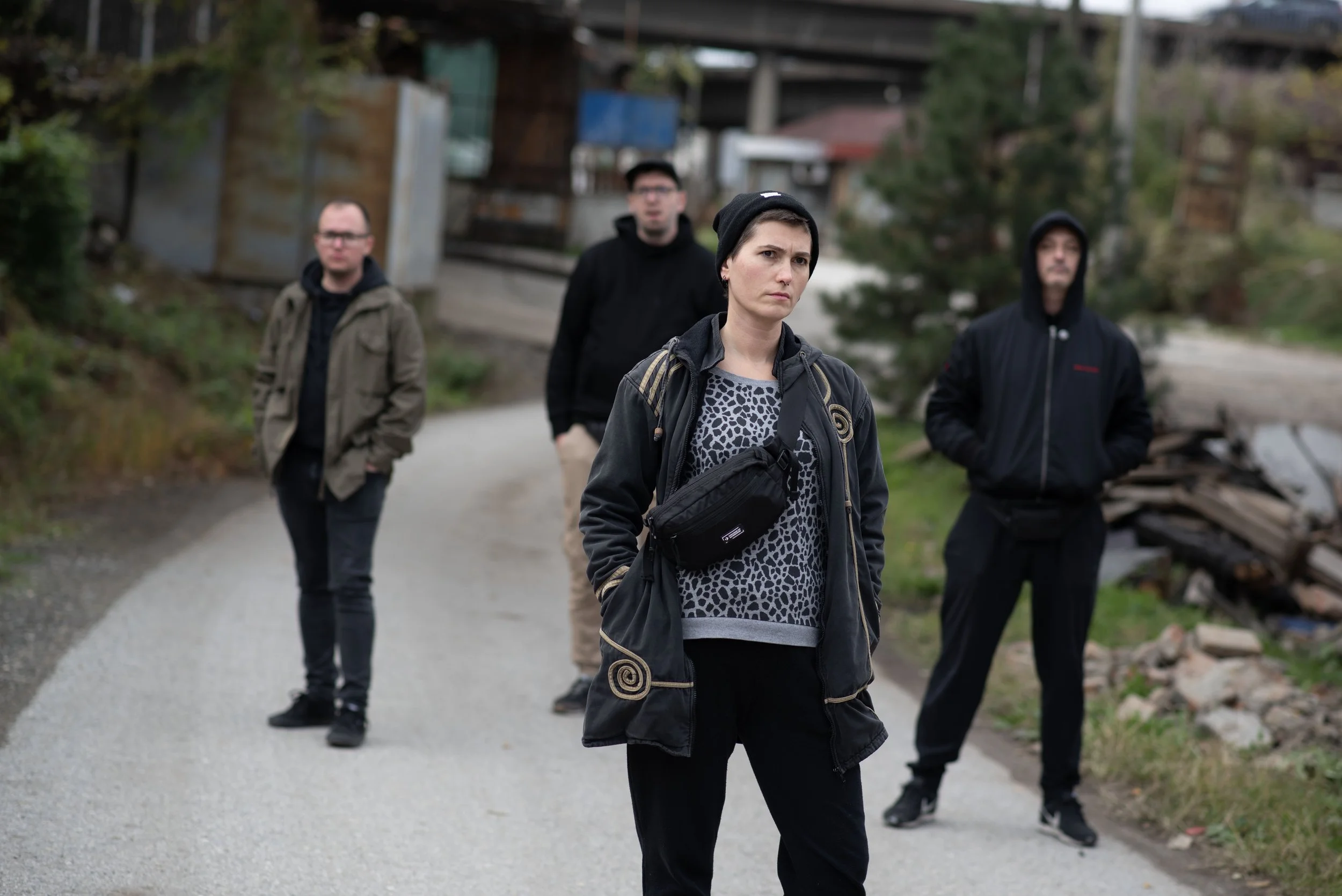 Four young adults walking outdoors on a gravel path in a rural area with trees and wooden structures in the background. The woman in the front is wearing a black jacket, a beanie, and a patterned shirt, looking serious. The other three men are behind her, dressed in casual jackets and hoodies.