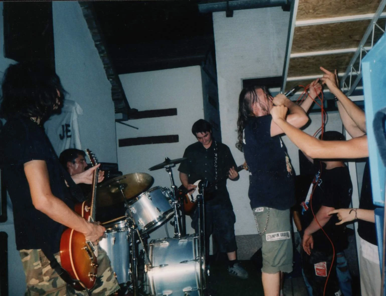 A band performing at a concert, with a guitarist, drummer, and vocalist, in an indoor venue with a high ceiling and exposed beams, and fans in the audience.