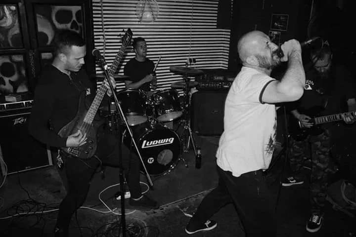 A band performing live in a music venue, featuring a vocalist with a microphone, two guitarists, and a drummer behind them.