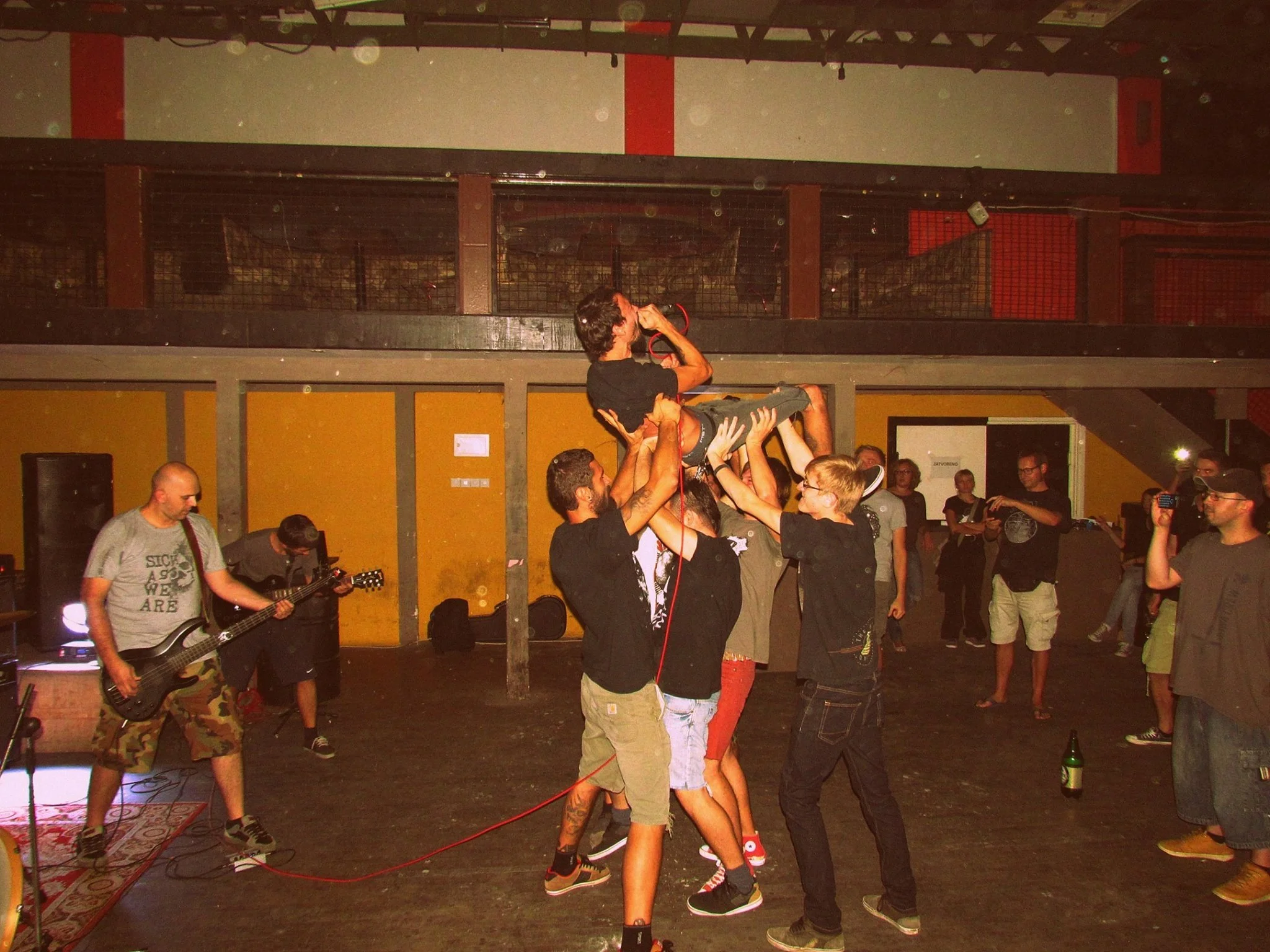 A group of people forming a circle at a concert or party, lifting a person in the air. The setting appears to be indoors with warm lighting and a stage area.
