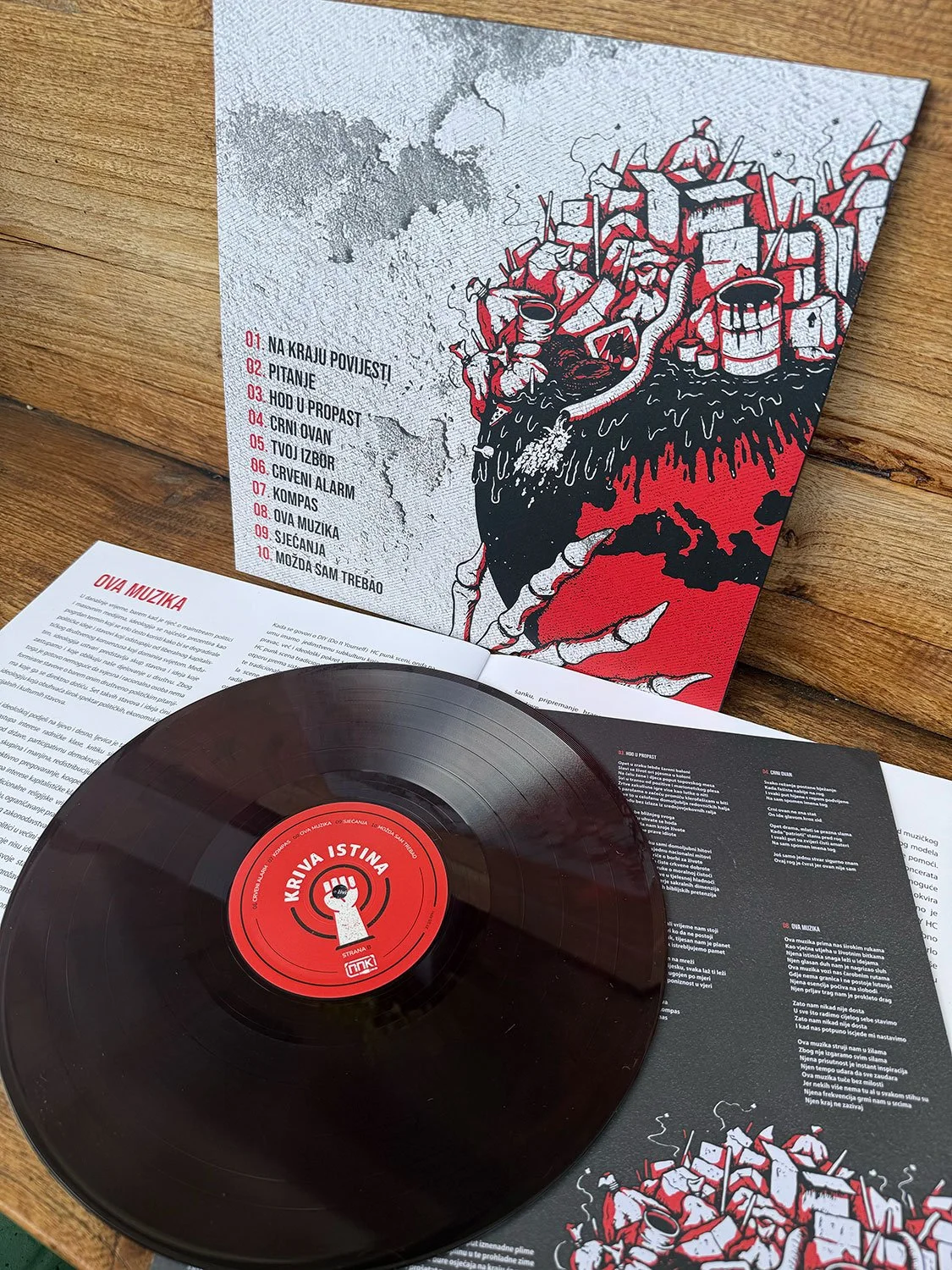 A vinyl record titled 'Kriva Istina' with its cover art showing a stylized black and red graphic of a heart with various objects on top, leaning against a wooden surface, with a booklet containing song lyrics and descriptions nearby.