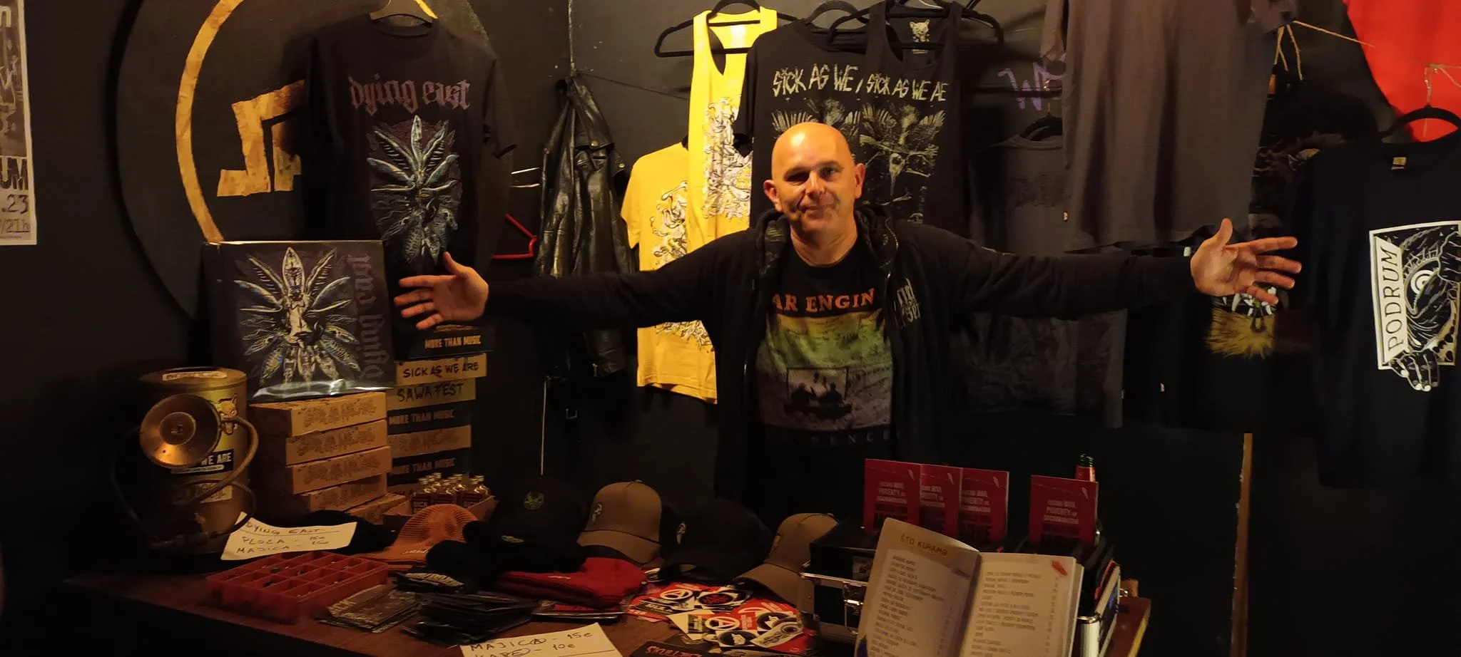 A man with arms outstretched standing behind a merchandise table at an event. The background features black clothing with psychedelic and rock band designs, including a shirt that reads "Fucked As We Are" and another that says "Sick As We Are." The t
