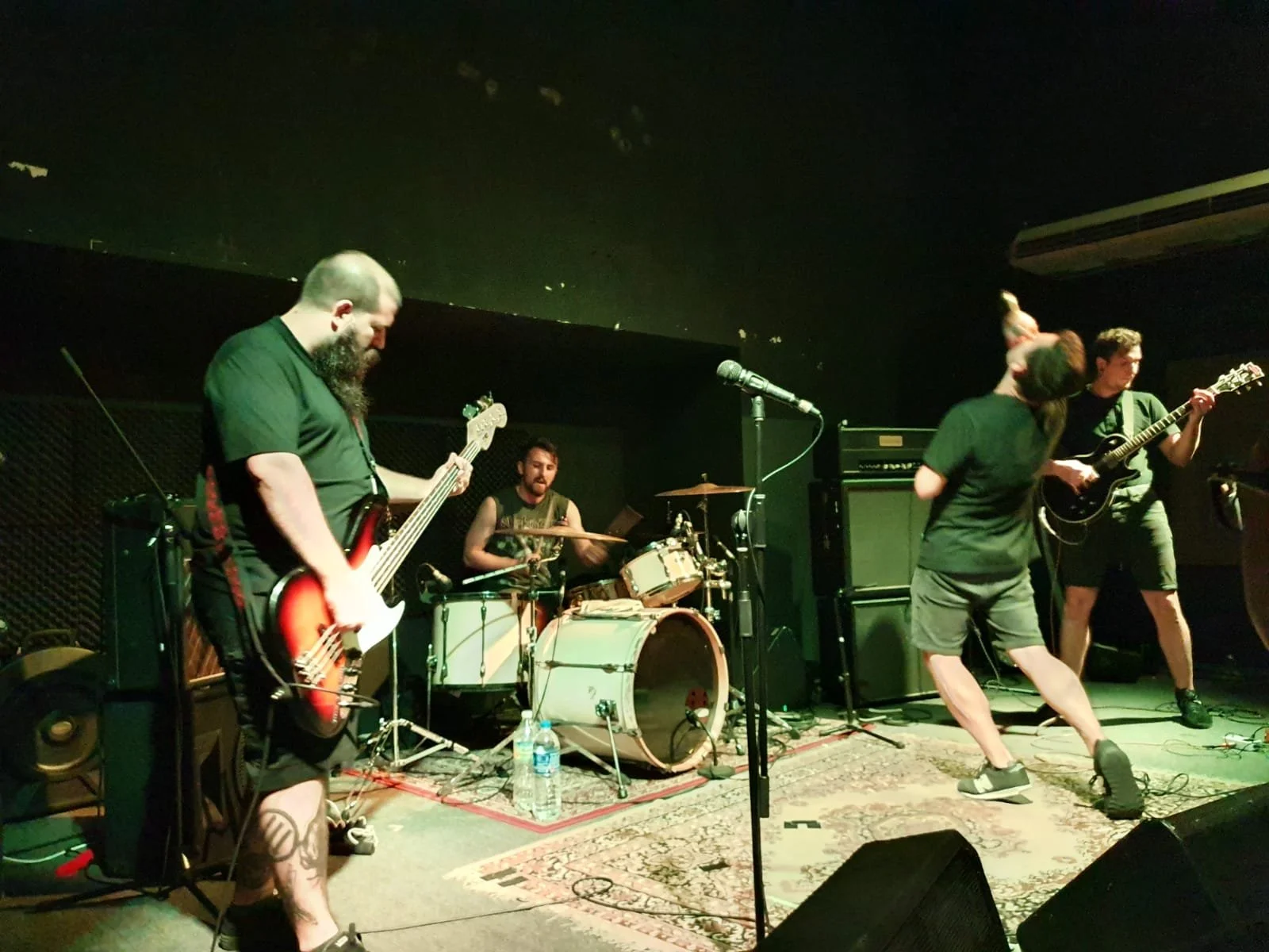 A live band performing on stage with three guitarists, a drummer, and a female singer. The stage has musical equipment, a rug, and speakers.