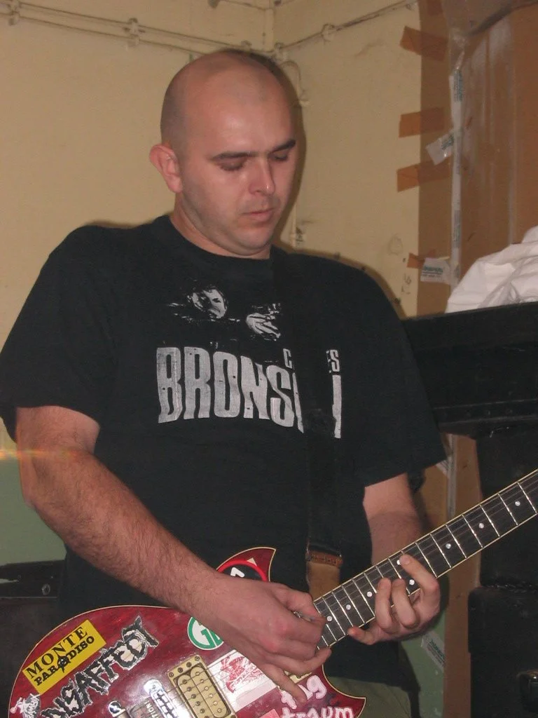 A man in a black t-shirt playing an electric guitar in a room of cardboard boxes.