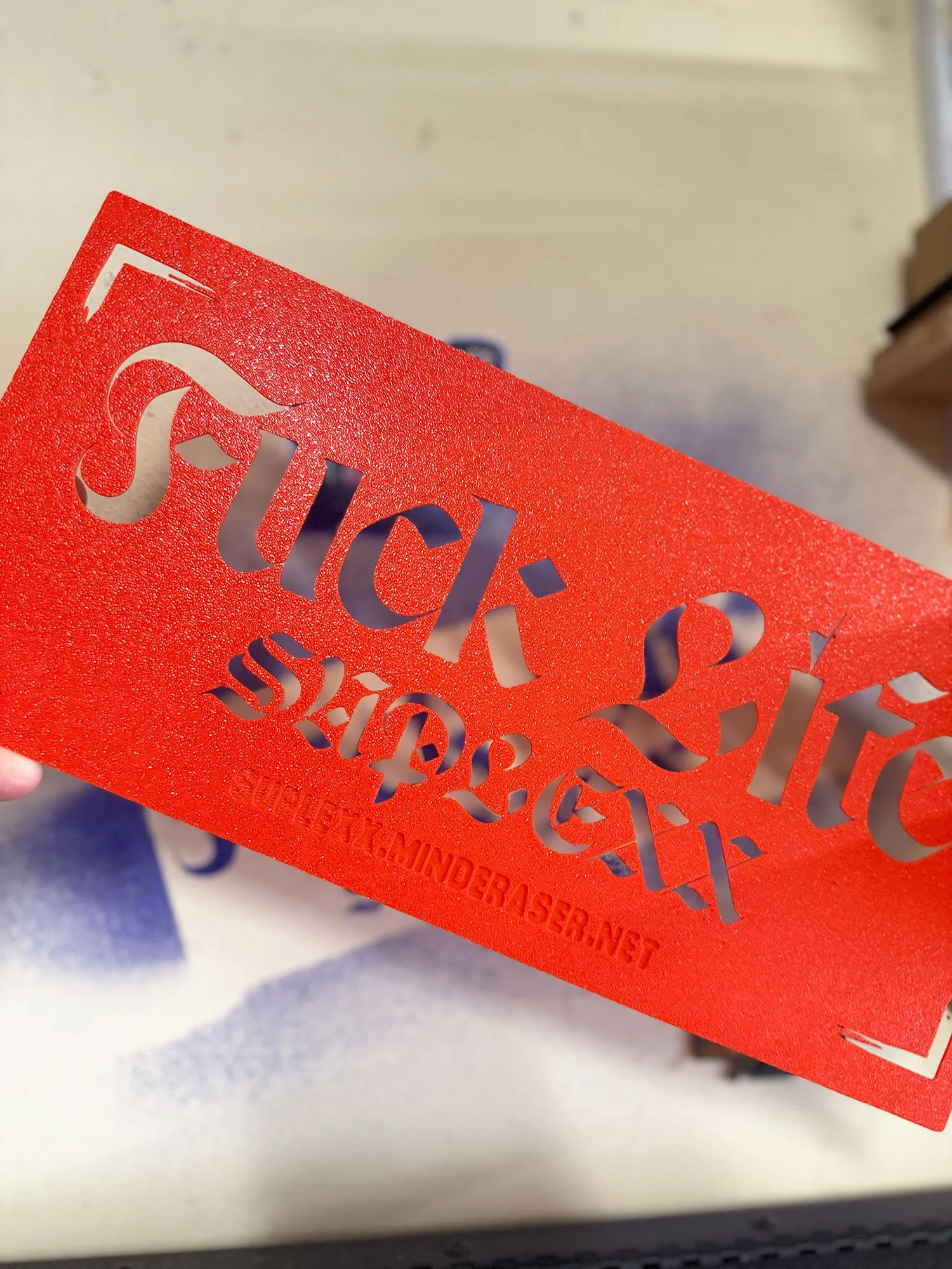 A red plastic stencil with cut-out text and design, held over a white surface.