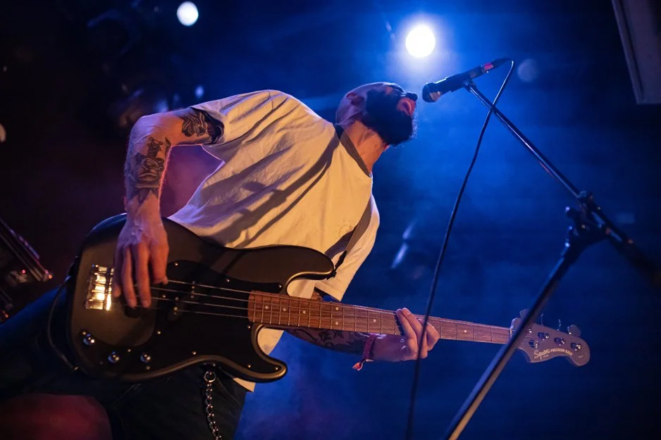 A bearded man wearing a white T-shirt and a cap, playing an electric bass guitar on stage with blue and purple lighting, microphone stand in front of him.