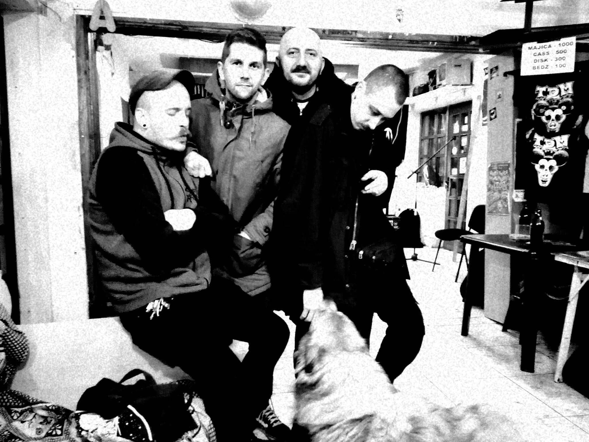 Four men sitting and standing inside a room with shelves and a whiteboard, one holding a dog, and a poster on the wall with gaming points listed.