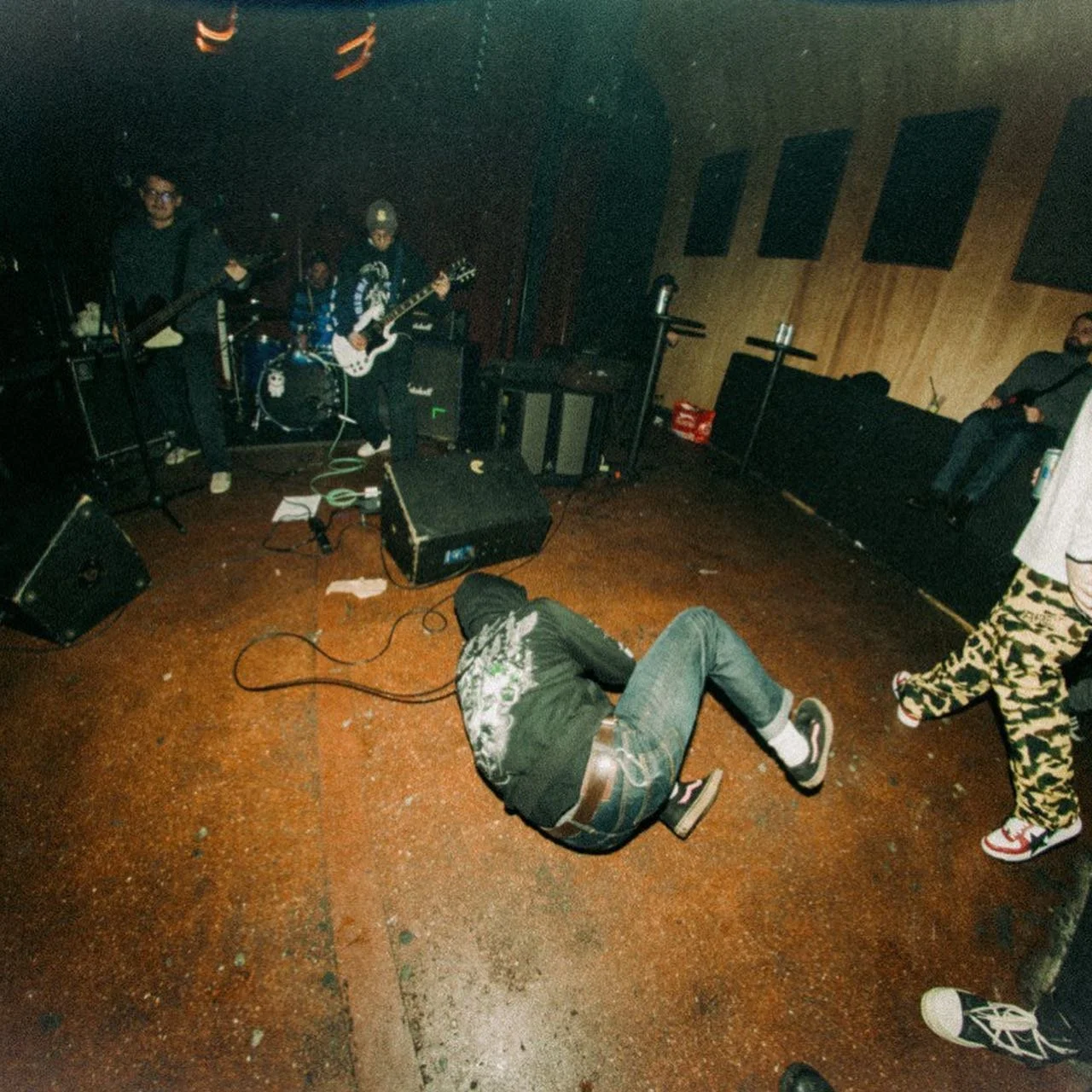 A band performing live music on stage with two guitarists and a drummer. Someone is lying on the floor in front of the stage, and a person wearing camouflage pants and red shoes is walking nearby. The room has wooden walls and a dark floor.