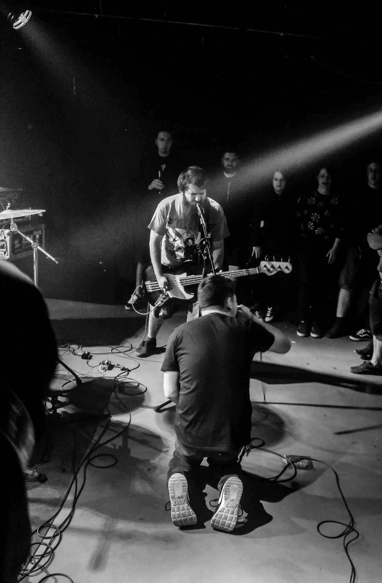 Black and white photo of a live music scene at a small venue. In the foreground, a man with a beard and glasses is kneeling on the floor, facing away, with several cables around him. Above him, a guitarist with shaggy hair and a graphic T-shirt is be