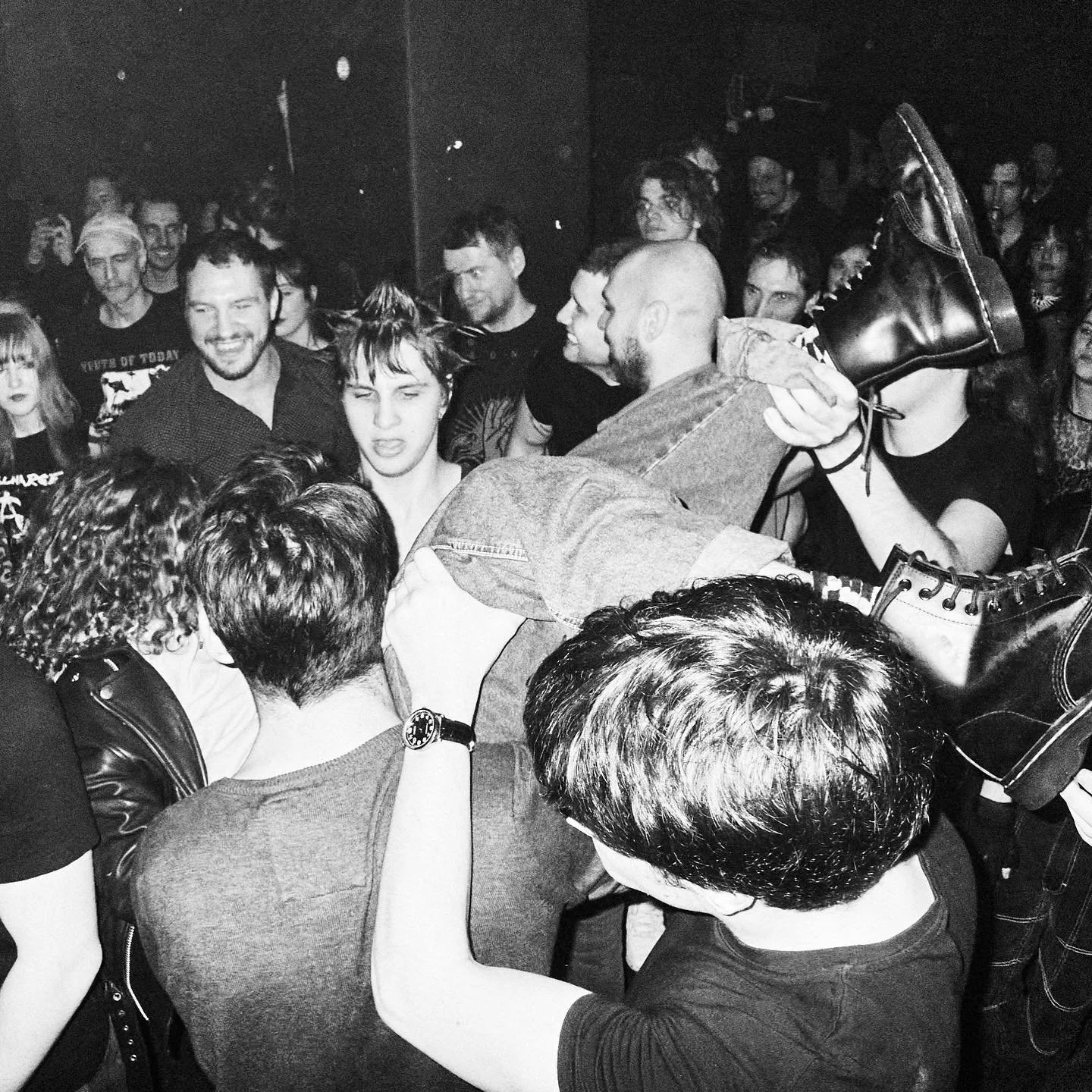A black and white photo showing a crowded concert scene with people crowd surfing. A person in the center is being held up by others, with their eyes closed and a slight grimace. Several individuals are surrounding the crowd surfer, some smiling and 