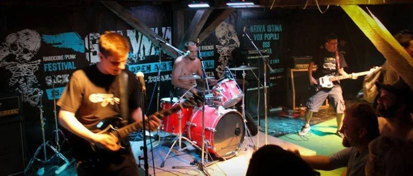 A band performing live on a stage with three members playing guitars and drums, with audience members watching in a dimly lit venue decorated with posters.