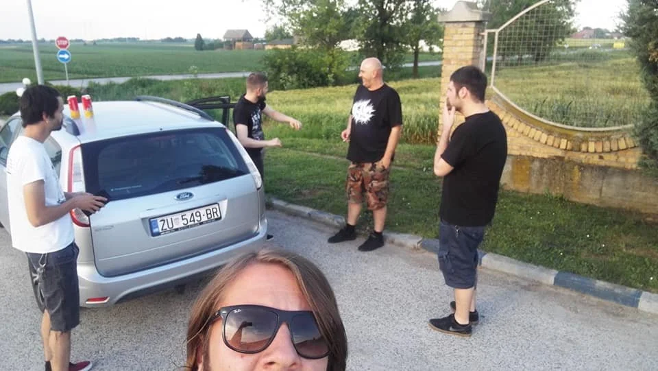 A man wearing sunglasses taking a selfie with four men in a rural outdoor setting. Three men are standing near a silver car, and one man is checking his phone. The scene appears casual with a grassy background and a stone wall.