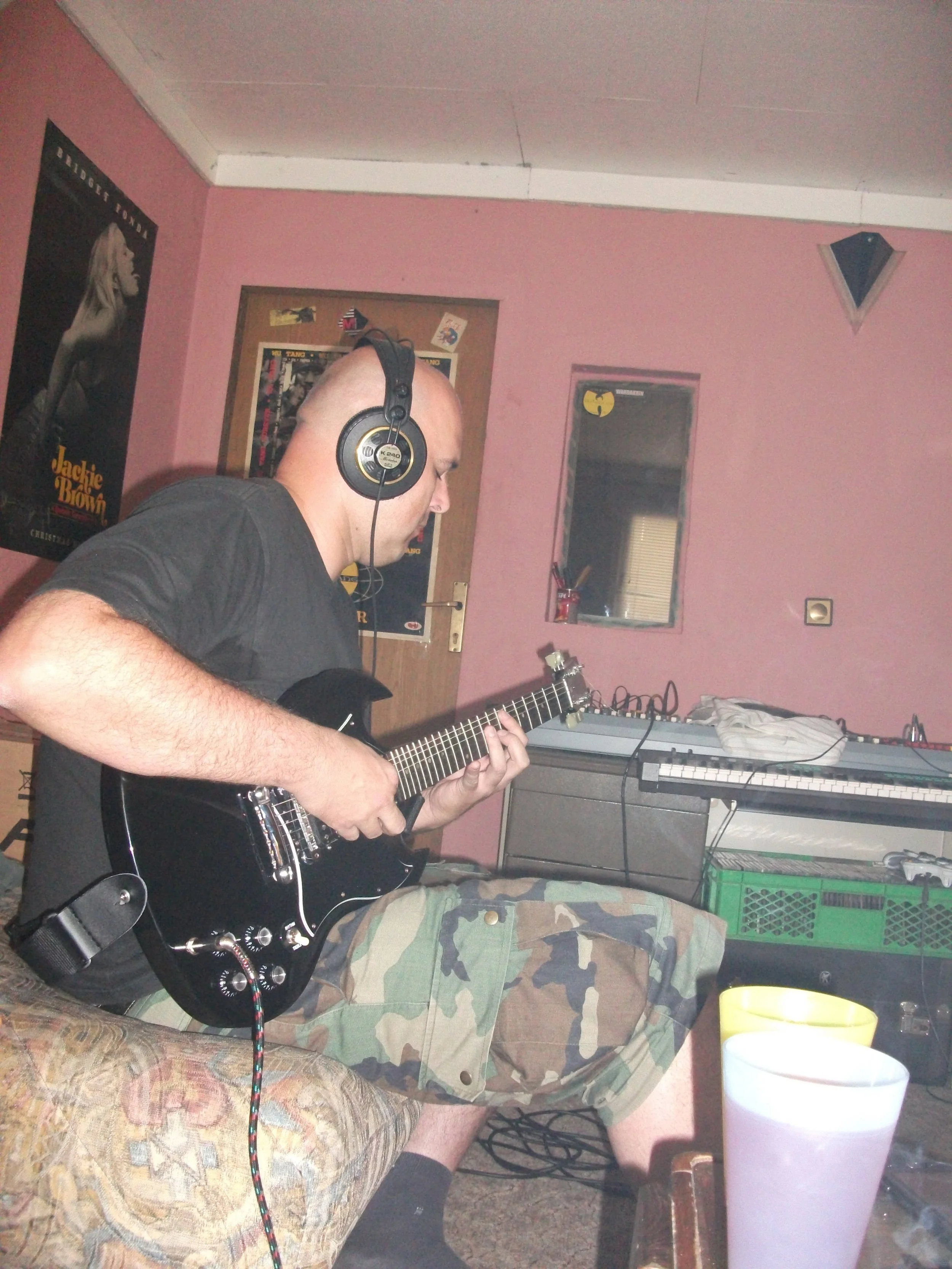 A man with a shaved head wearing headphones and a black shirt playing an electric guitar while sitting on a couch in a room with pink walls, posters, and a window.