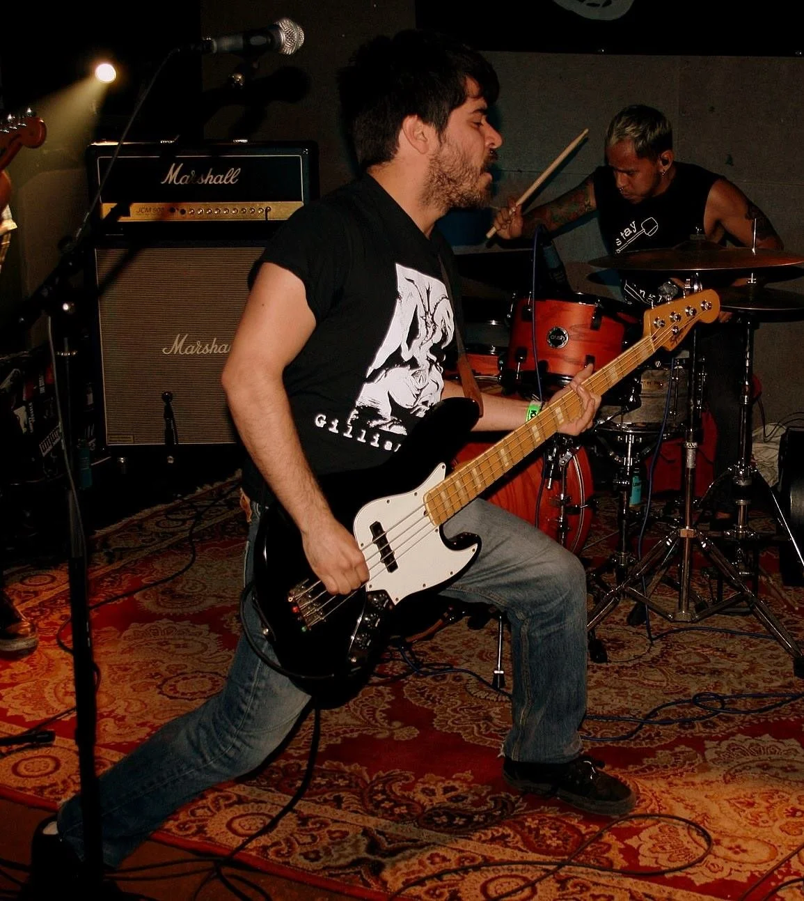 A musician playing a bass guitar on stage with a drummer in the background, Marshall amplifiers visible, performing in a dimly lit venue.
