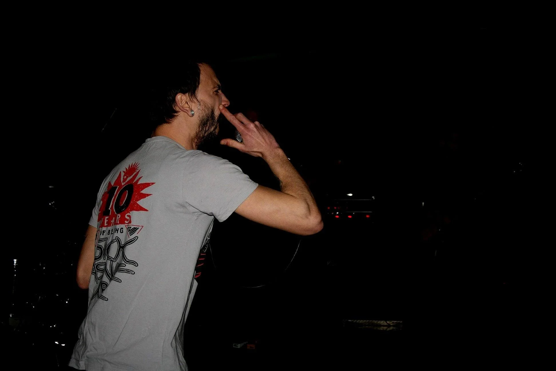 A man with dark hair and a beard, wearing a gray T-shirt with a graphic print on the back, is holding his nose with his right hand against a dark background.
