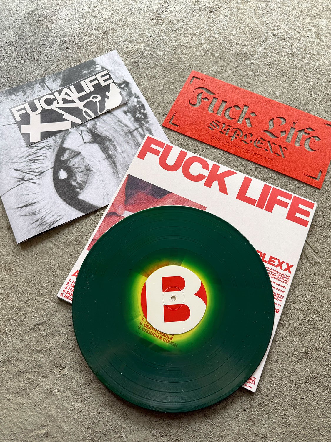 Green vinyl record with a white and red label on a background of red and black scanned music album covers and a red sticker, all on a concrete surface.
