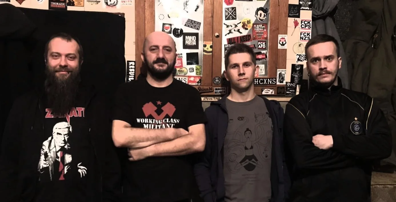 Four men standing in front of a wall covered with stickers and posters, with arms crossed or relaxed.