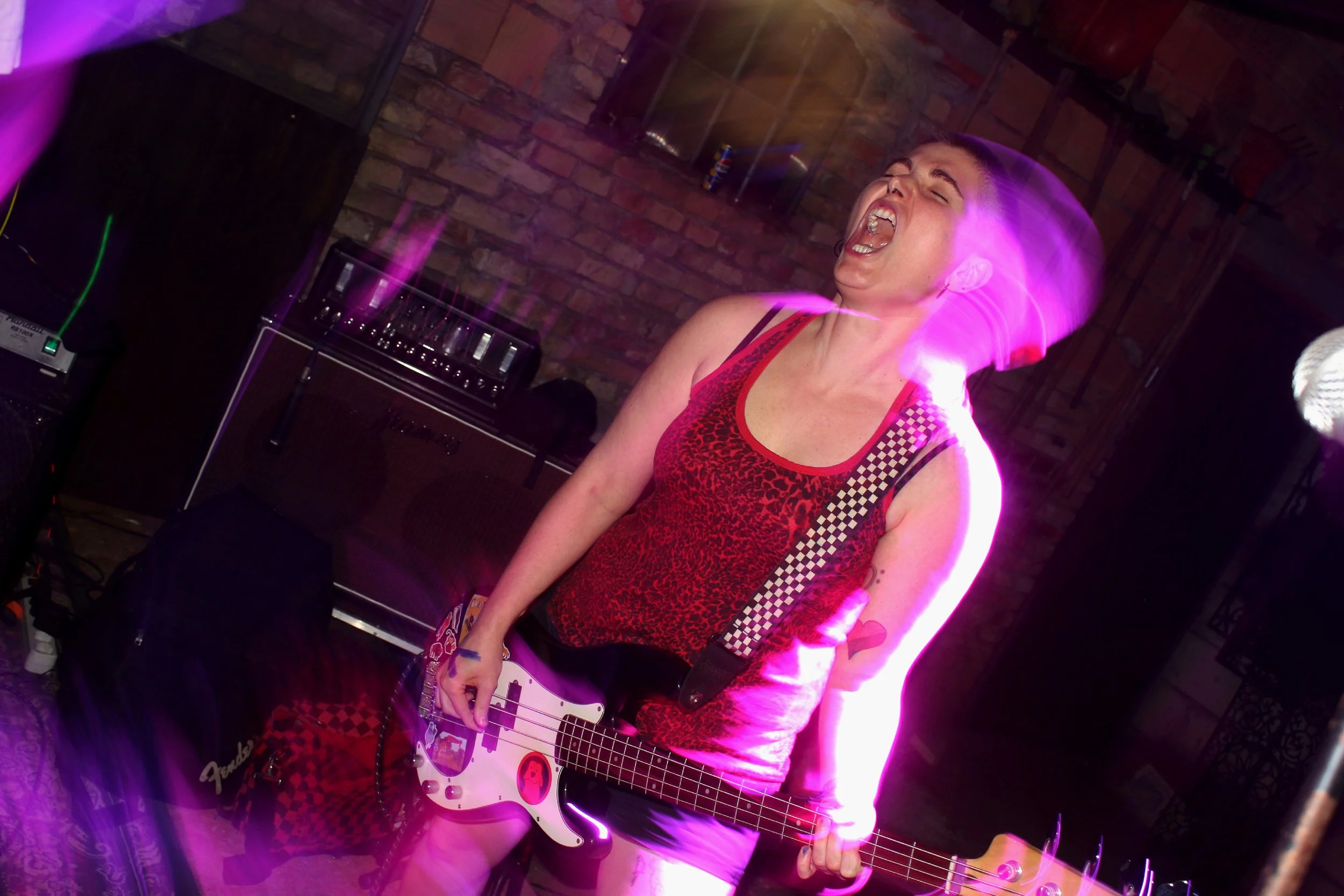 A person passionately singing into a microphone while playing an electric guitar, with vibrant pink and purple lights and a brick wall background.