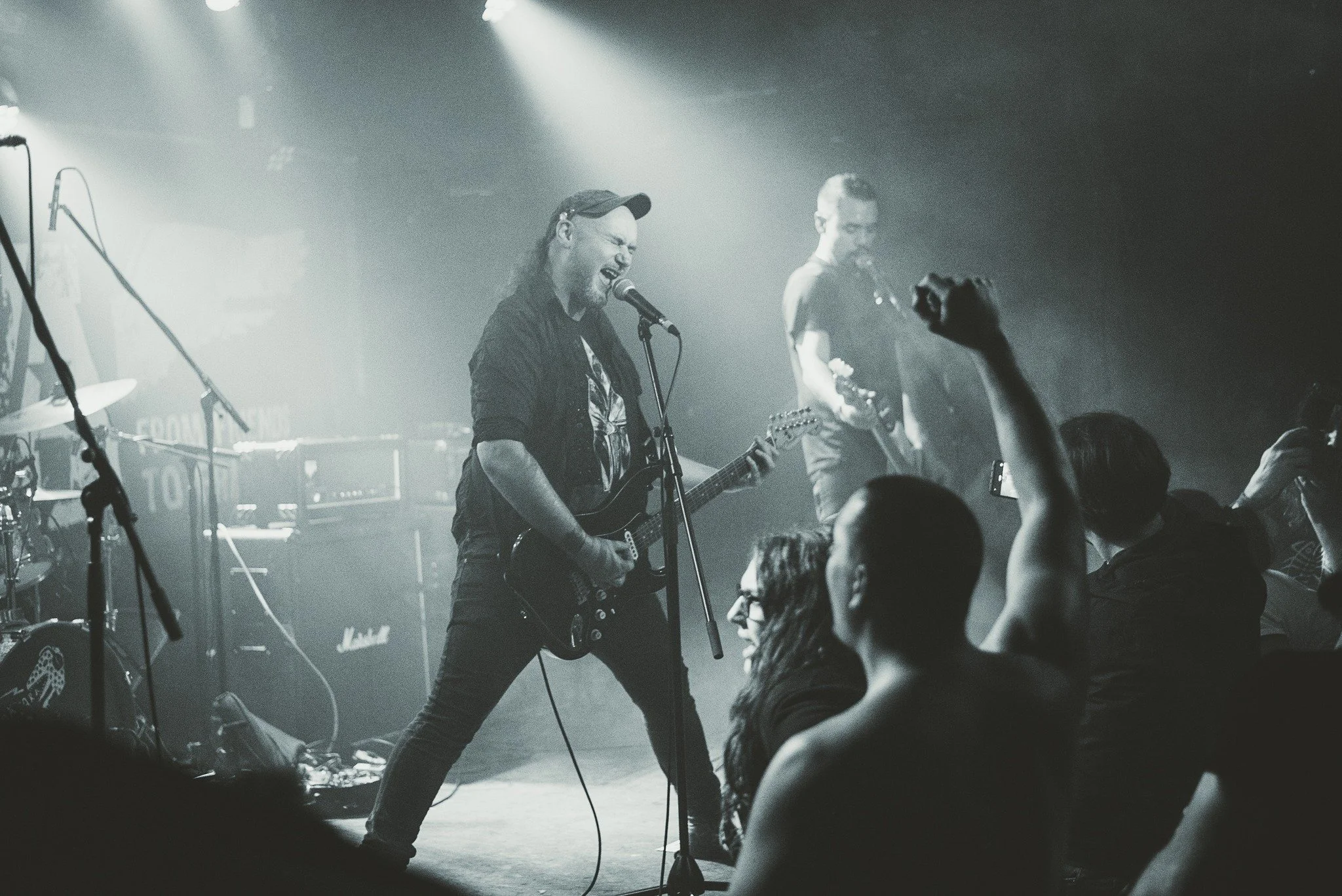 A black-and-white photo of a live band performance on stage, with a lead singer playing an electric guitar and singing into a microphone, and another band member in the background playing an instrument, with an audience in front raising their hands.
