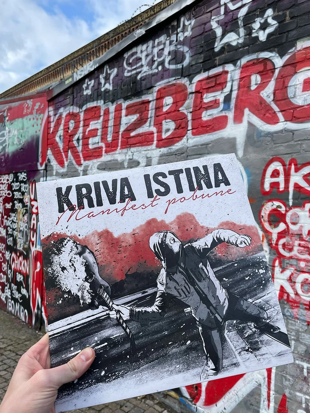 A hand holding a poster with an illustration of a person breaking a coffee pot with a spray of coffee, labeled 'KRIVA ISTINA' and 'Manifest pobune,' in front of a graffiti-covered brick wall with the word 'KREUZBERG' painted in red.