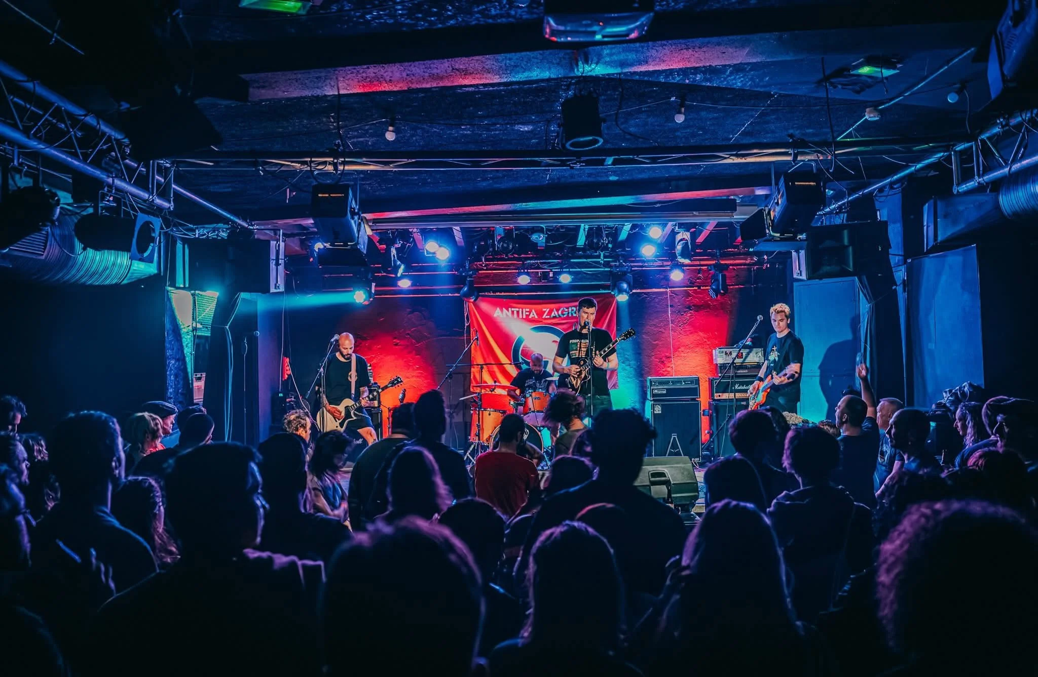A live band performing on stage with four members, one playing drums, two with guitars, and one with a keyboard, in front of an audience in a music venue with colorful lighting and a red banner that reads 'ANTIFA ZAGREB' at the back.
