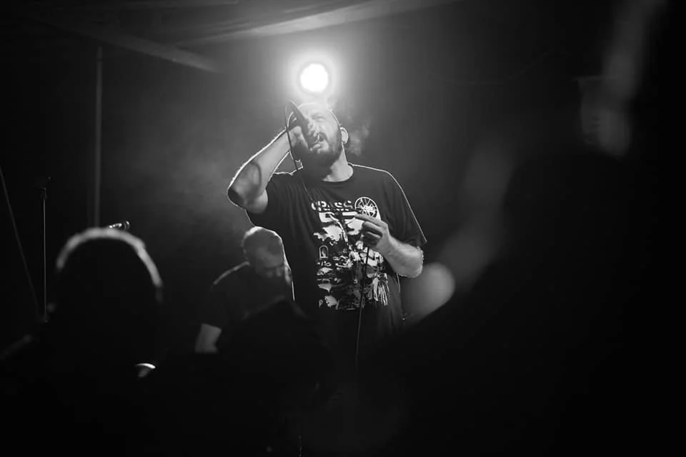 A man singing into a microphone on stage, illuminated by a bright light behind him, with a band member playing guitar in the background, in a dark concert setting.
