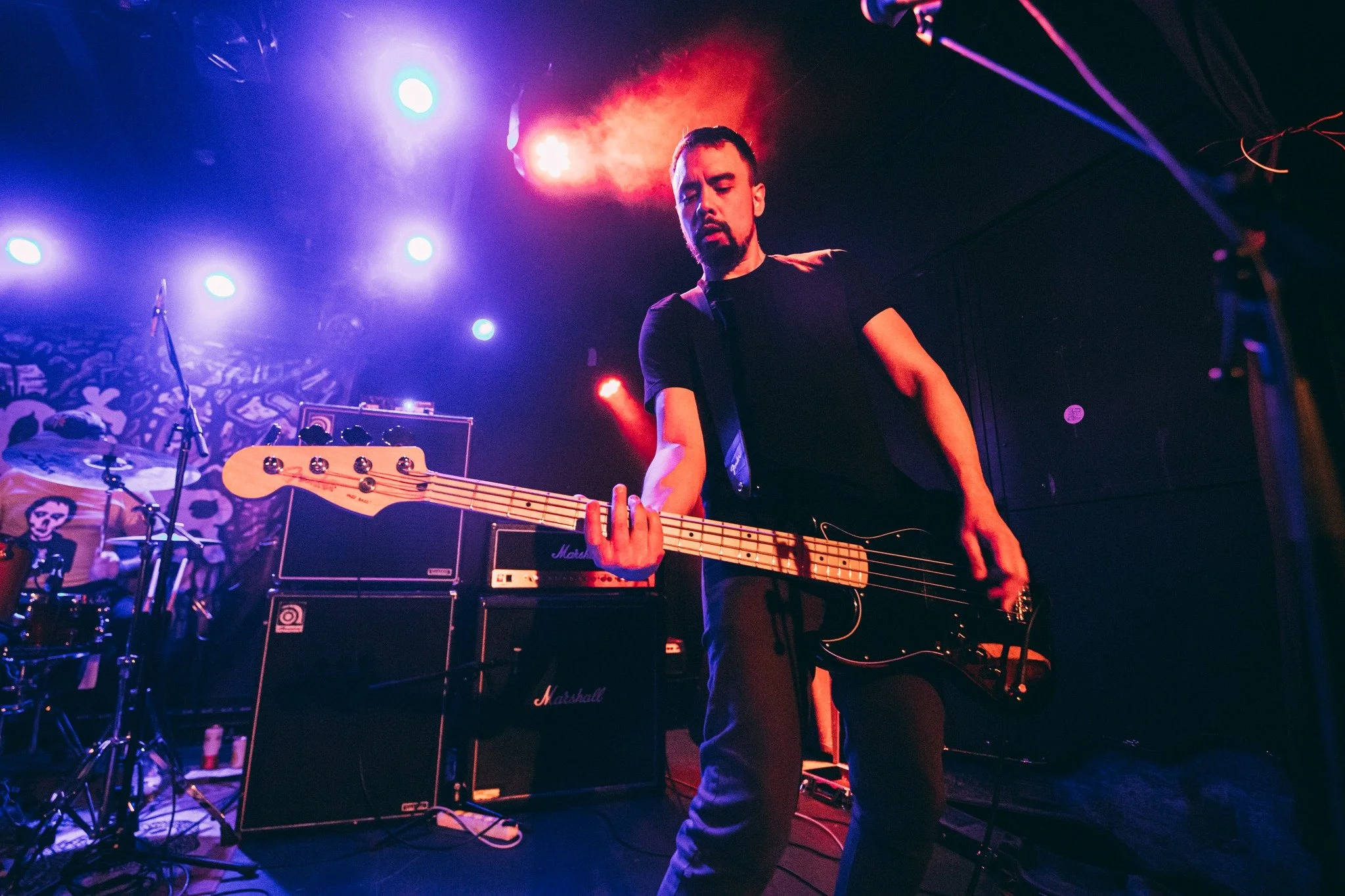 A man with a beard playing a bass guitar on stage, illuminated by colorful stage lights, with musical equipment such as amplifiers and a drum set around him.