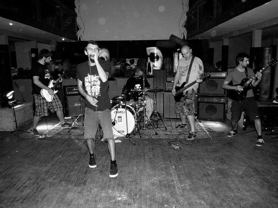 A band of five young men playing instruments and singing on a stage in a dimly lit venue.