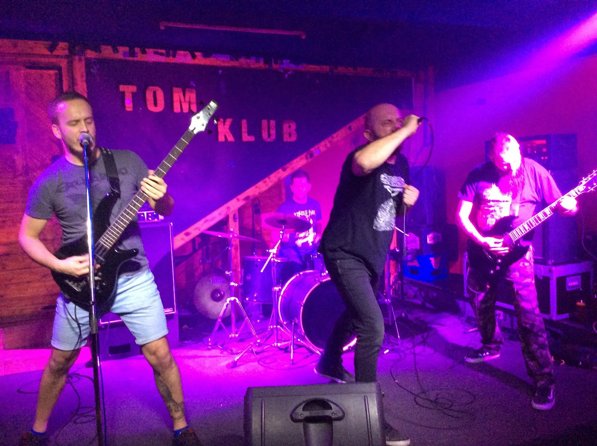 A band performing live on stage at a club with purple lighting, with a guitarist, a vocalist with a microphone, a drummer in the background, and another guitarist on the right. The stage has a sign in the background that says 'TOM' and 'KLUB'.