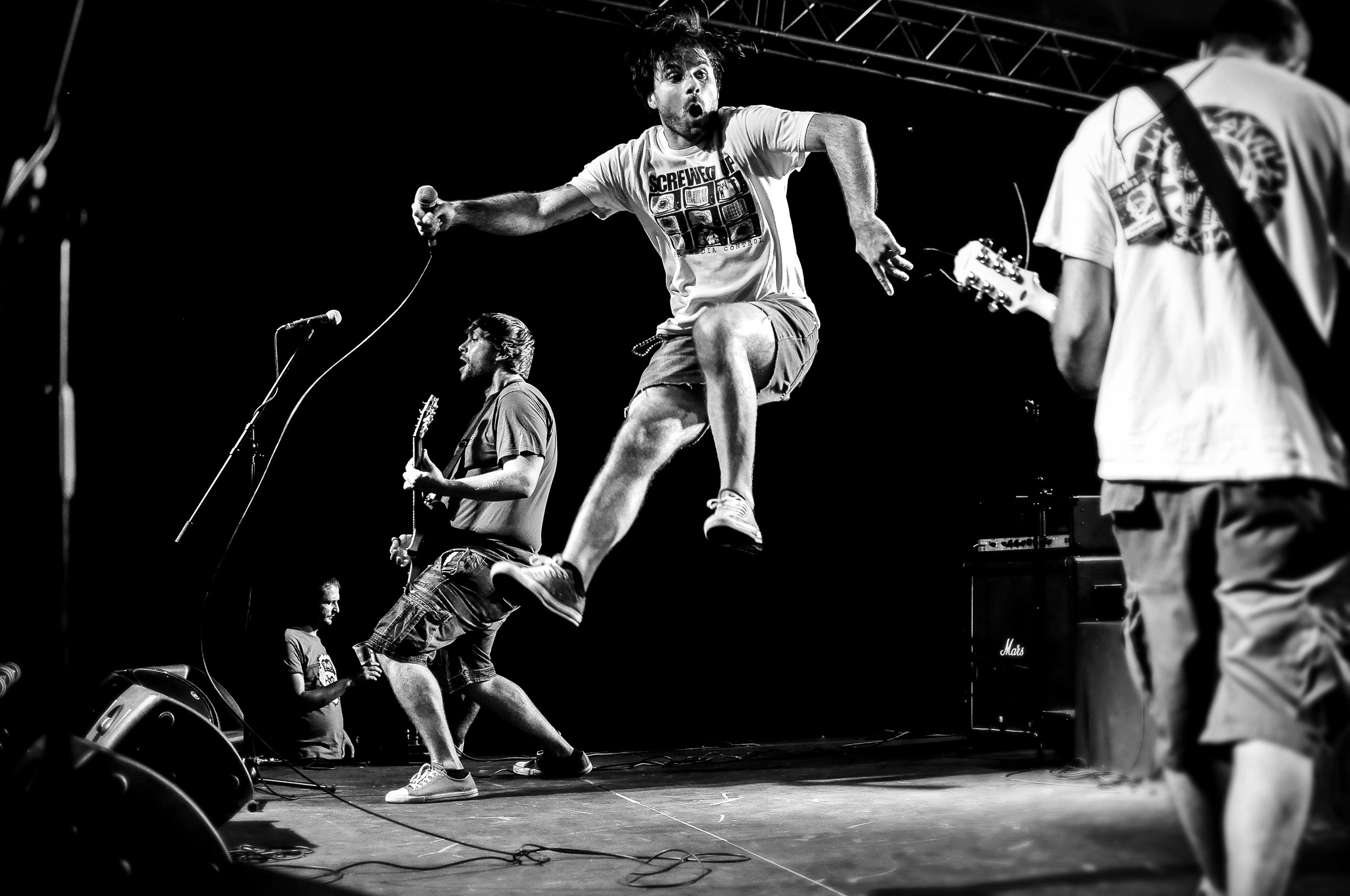 Black and white photo of a band performing on stage, with a singer jumping in the air holding a microphone. Two guitarists are playing, one in the background and one in the foreground facing away. There are large speakers at the front of the stage an