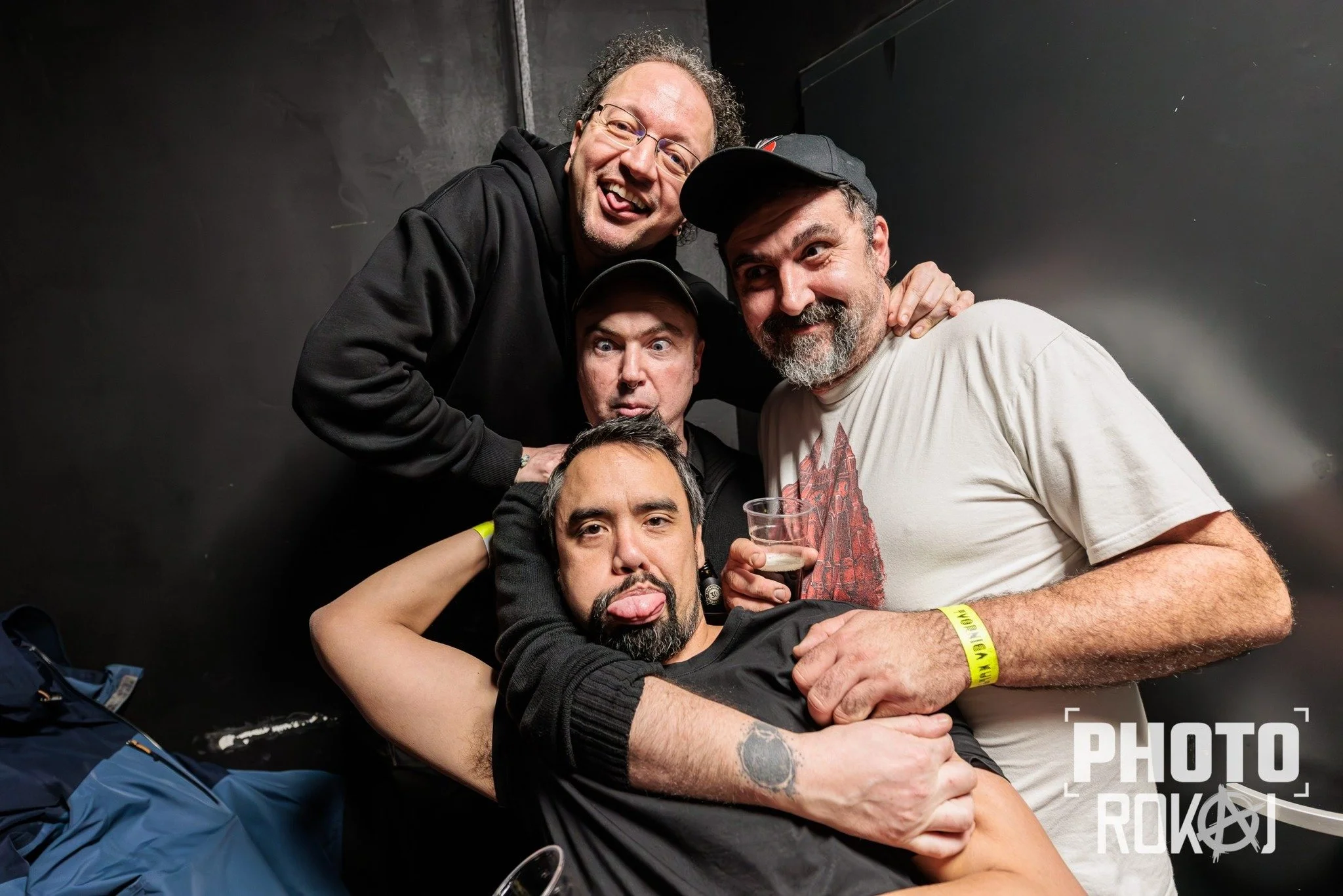 A group of five men from band LOS PRIMJER smiling and posing closely together in a dark indoor setting, with some holding drinks and making playful faces.