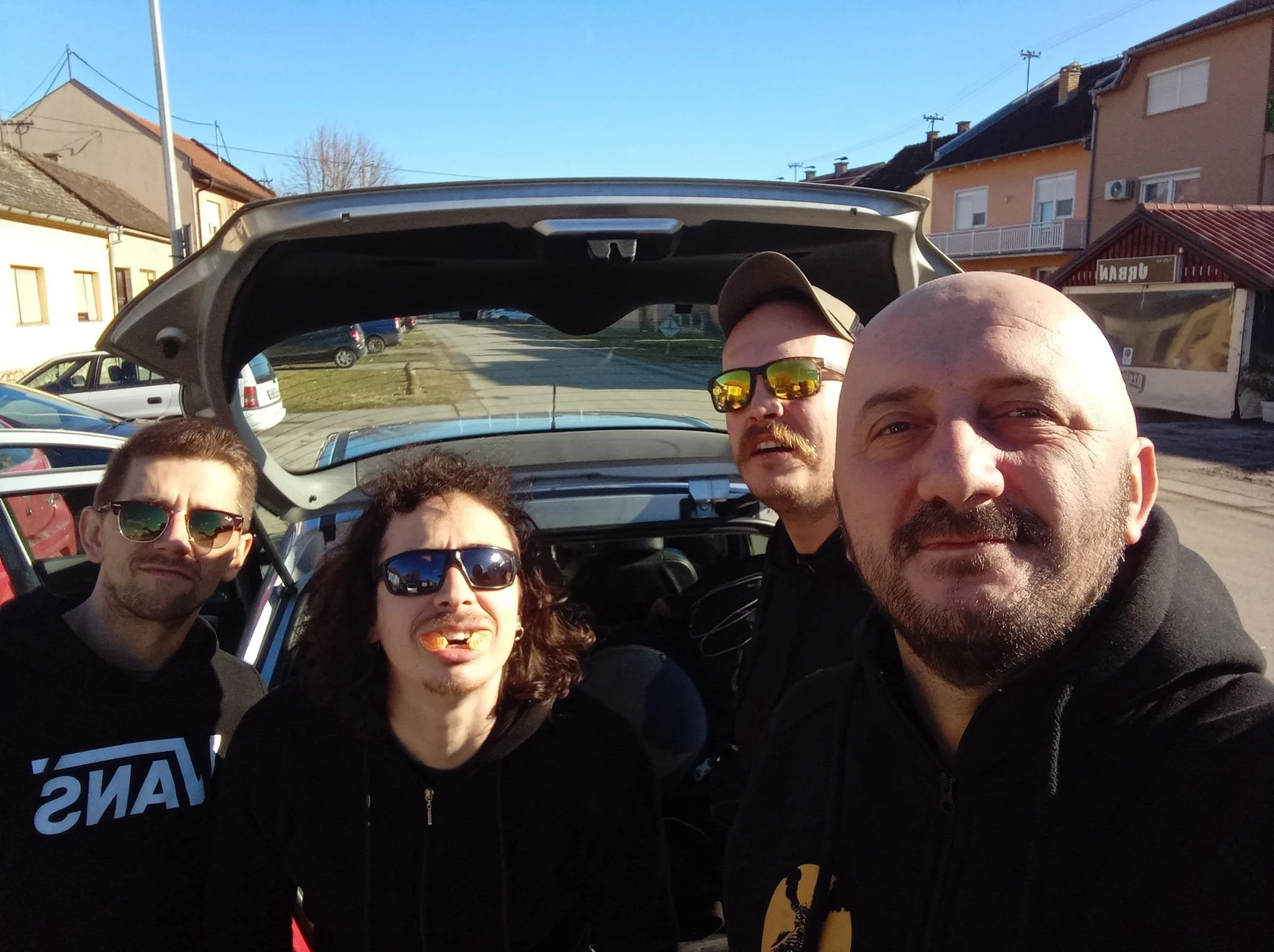 Group of five people taking a selfie outdoors during daytime, standing in front of an open car trunk, with residential houses and clear blue sky in the background.