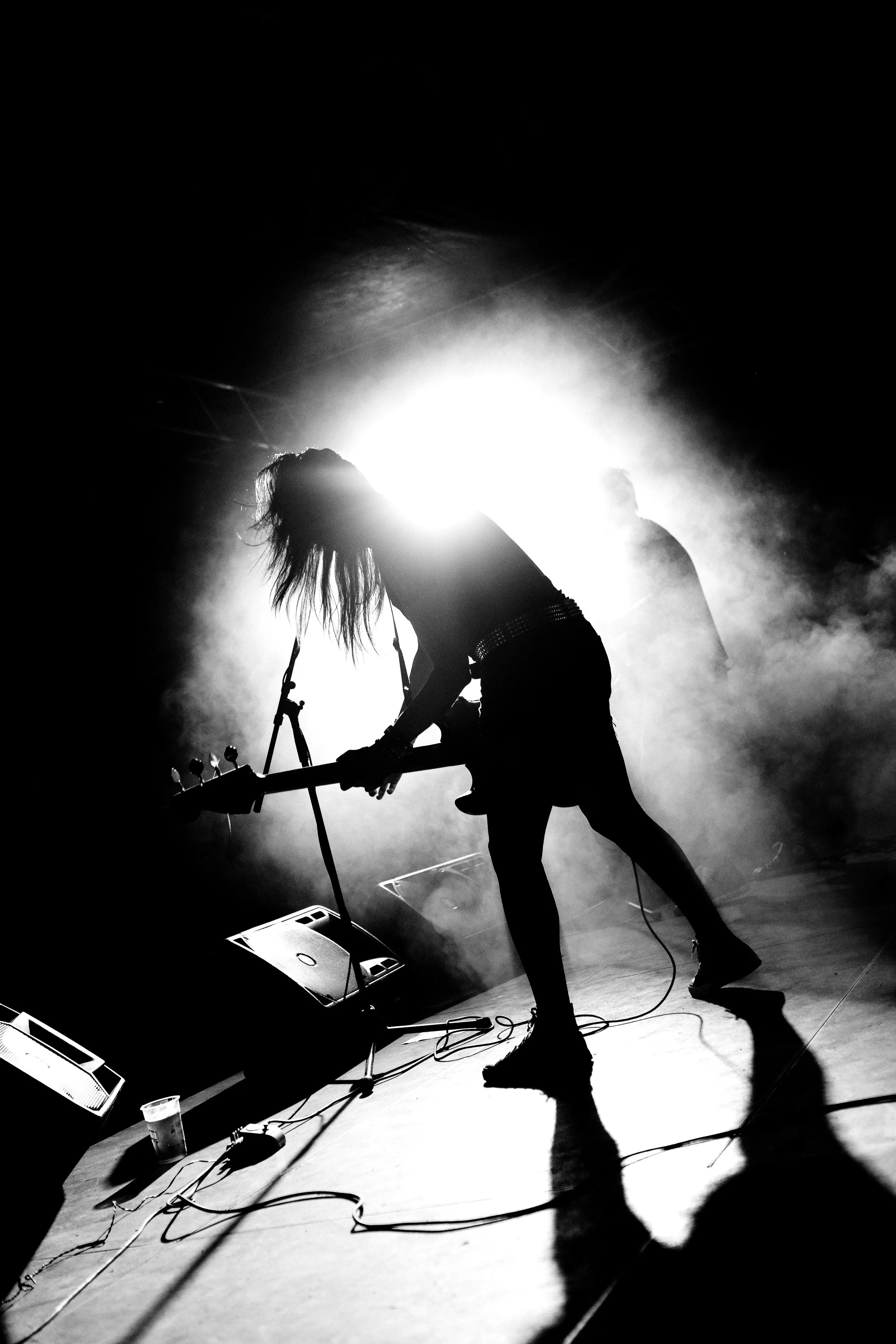 A black and white photo of a musician playing electric guitar on stage, backlit by bright lights, with a foggy atmosphere, and a person in the background. Band name: Concrete Worms