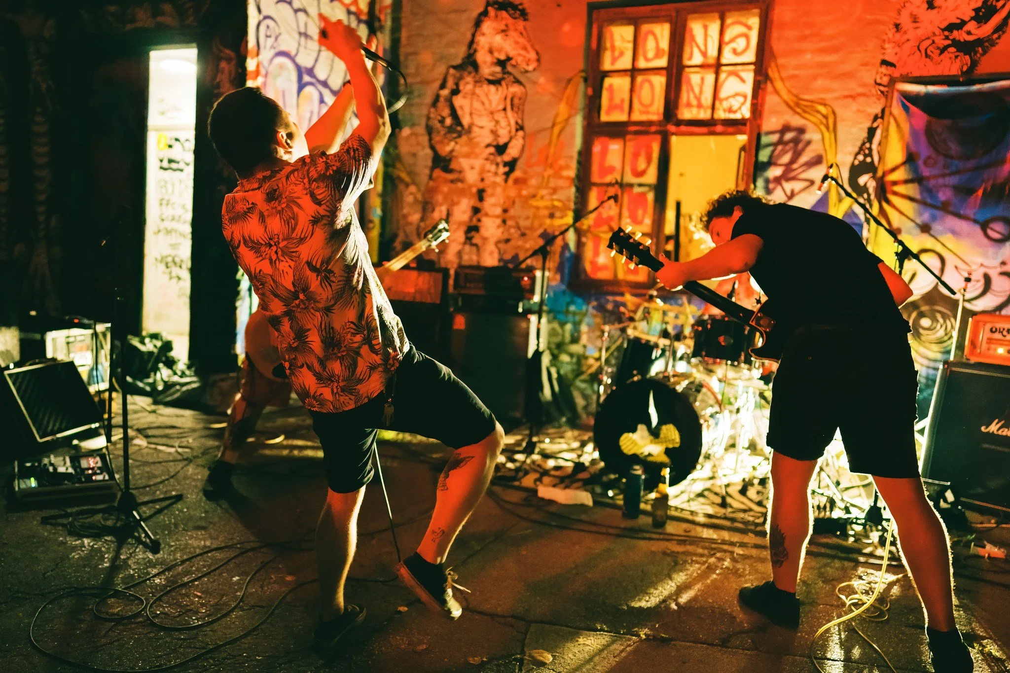 Musicians performing live on stage at a vibrant, graffiti-covered venue with colorful lighting, including a person in a floral shirt holding a microphone and another person playing an electric guitar.