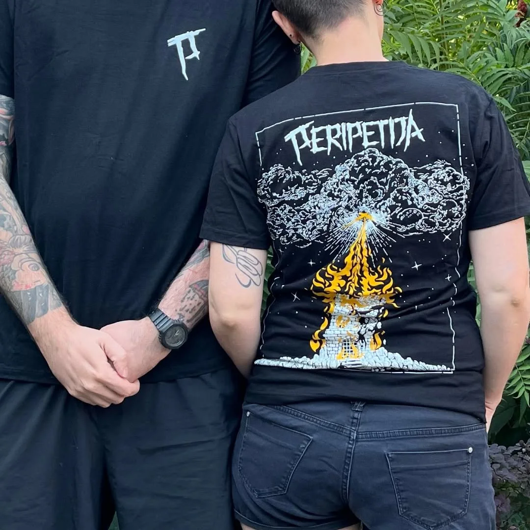 Two people standing outdoors, one with visible tattoos on their arm, wearing a black T-shirt with a small design, and the other wearing a black T-shirt with a large graphic and the word 'PERIPETIJA' on the back, surrounded by green foliage.