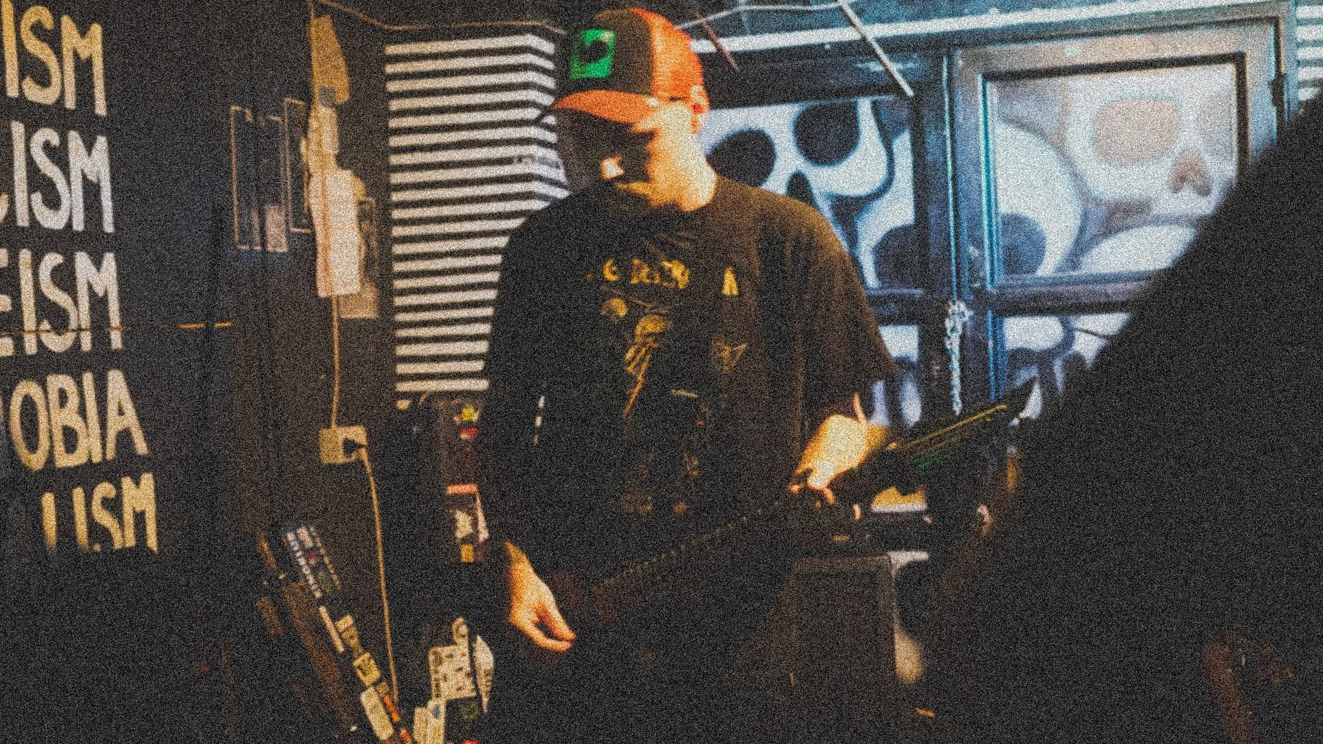 A person wearing a black t-shirt and a red baseball cap with orange brim holding a firearm in a dimly lit, graffiti-covered room with skull artwork on the wall.