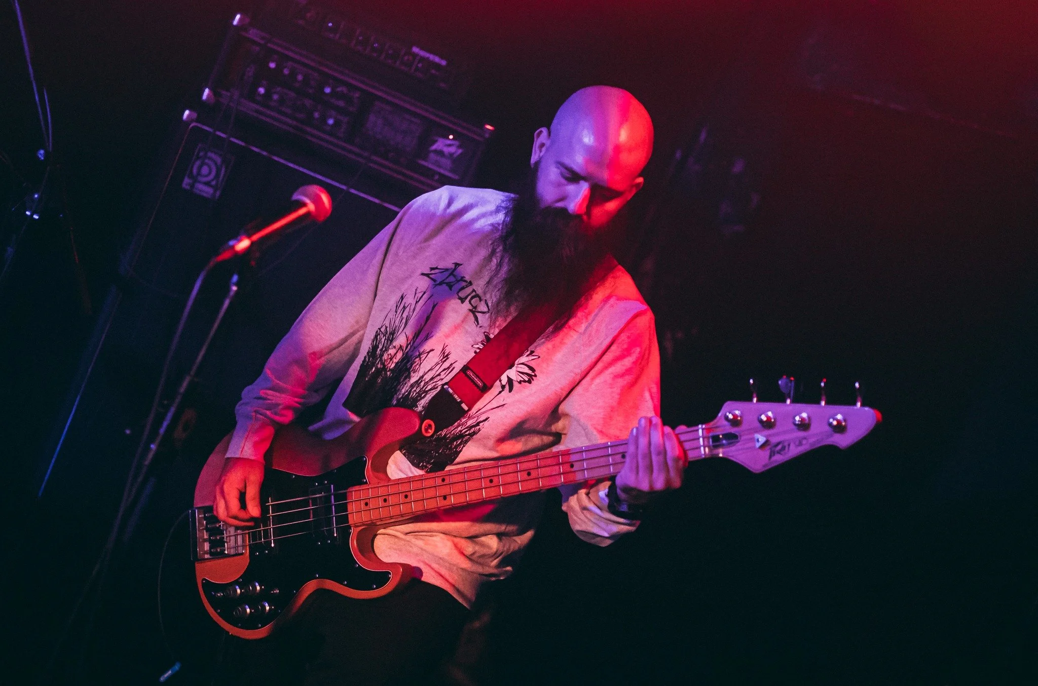 A man with a bald head and a long beard playing an electric bass guitar on a dark stage with purple lighting. There are sound equipment and a microphone nearby.