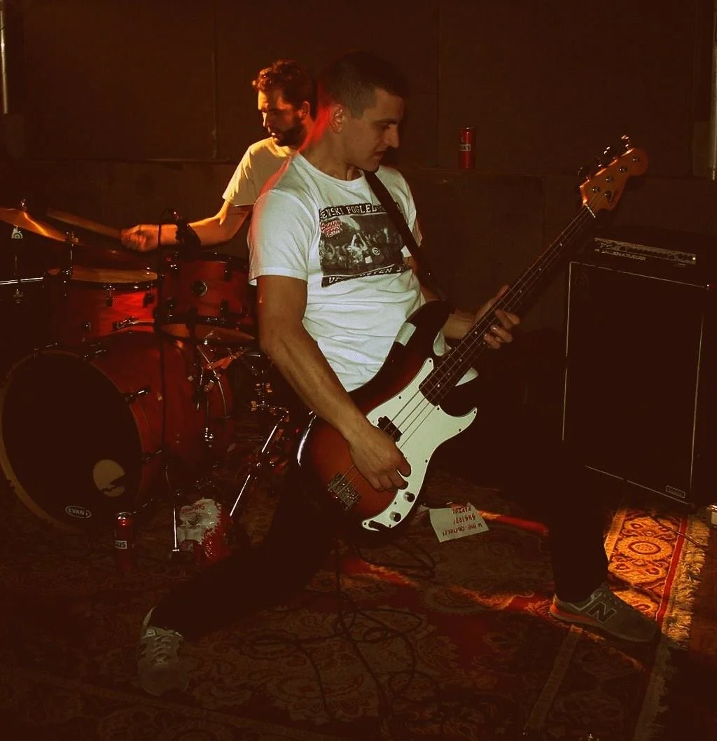 Two musicians performing on stage: one playing drums in the background and the other playing electric bass guitar in the foreground, with a dark background and stage lighting.