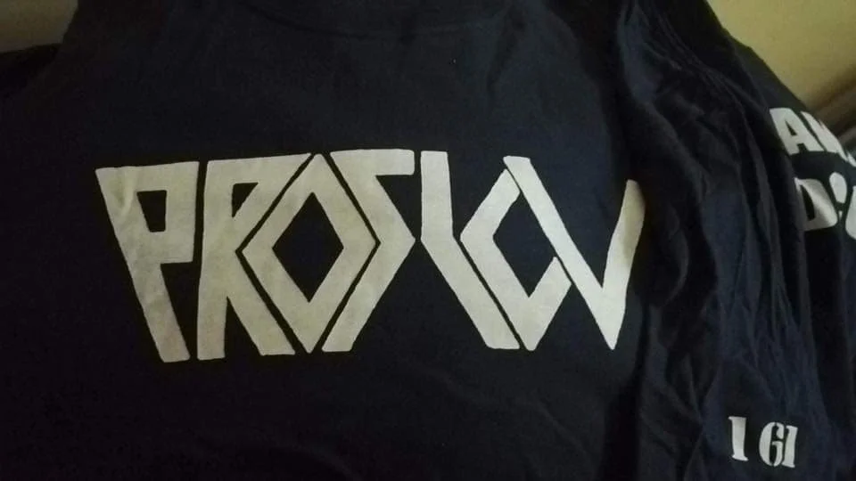 A person wearing a black shirt with white text that says 'PROSLOV' and a black jacket with some letters visible.