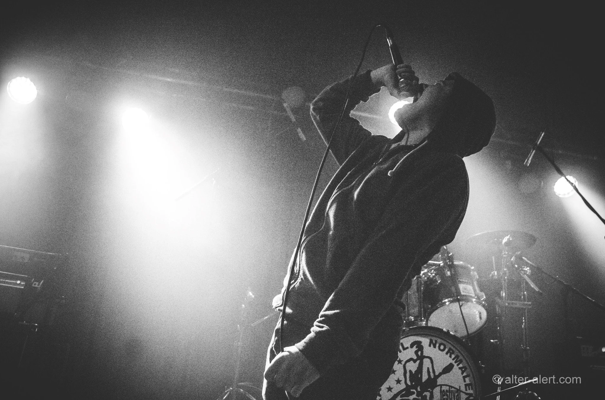 A silhouette of a person on stage holding a microphone and singing, with bright stage lights behind them, and a drum set visible in the background.
