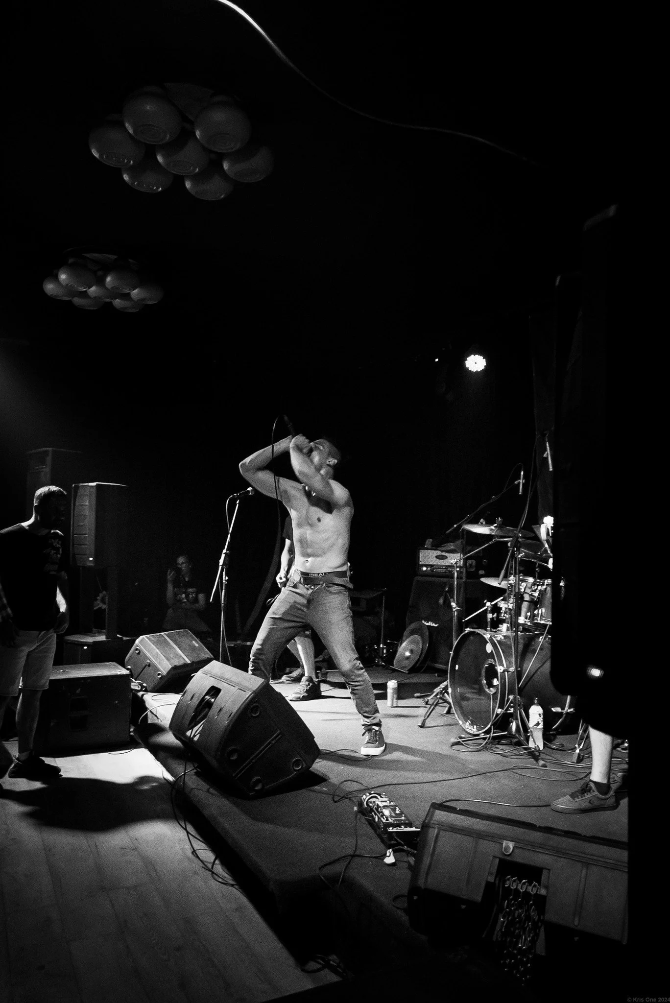 A musician performing shirtless on stage with musical equipment and band members visible in the background, under stage lighting.